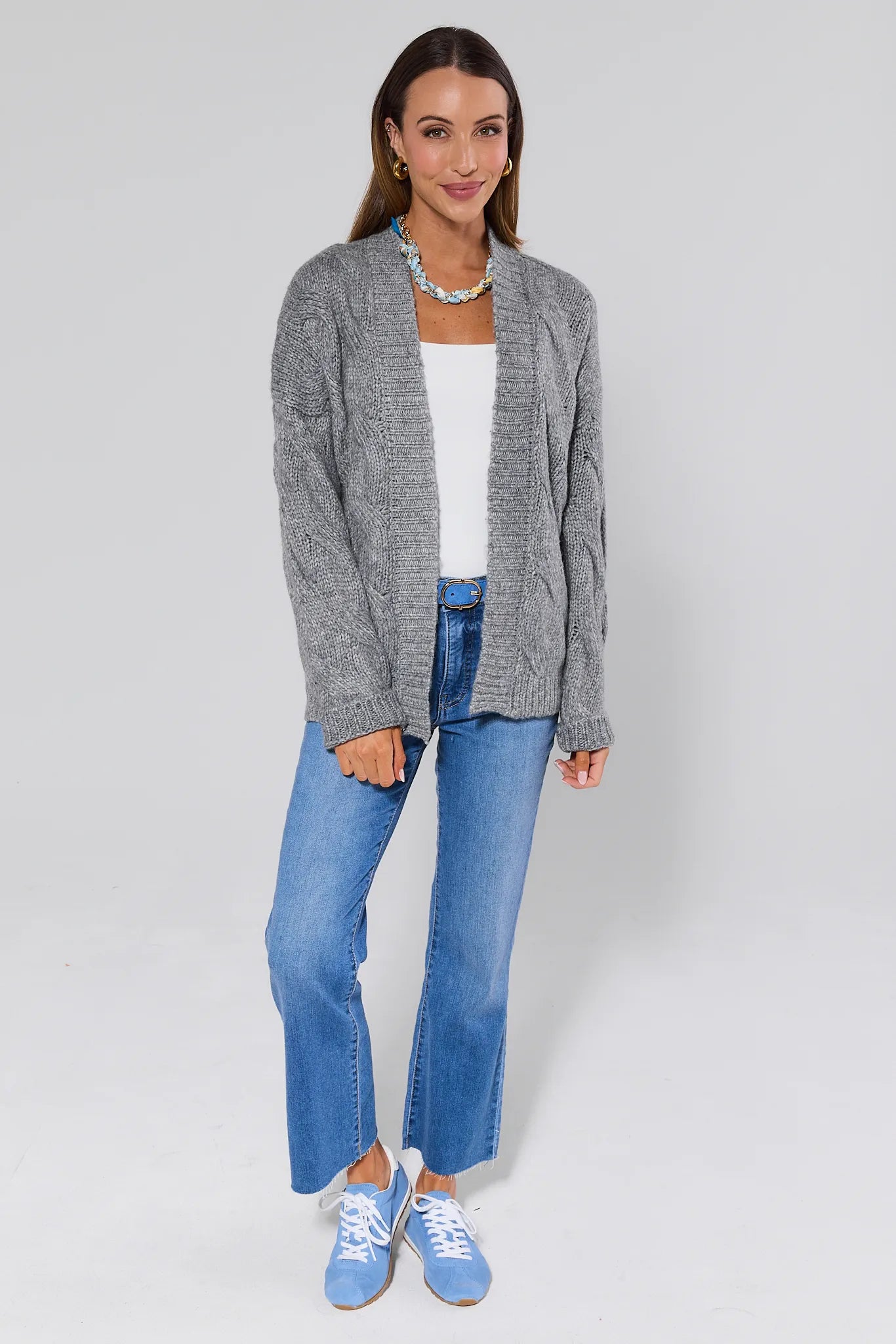 Woman wearing a Frankie Gray Cable Knit Cardigan over a white top and blue jeans and sneakers on a light gray background