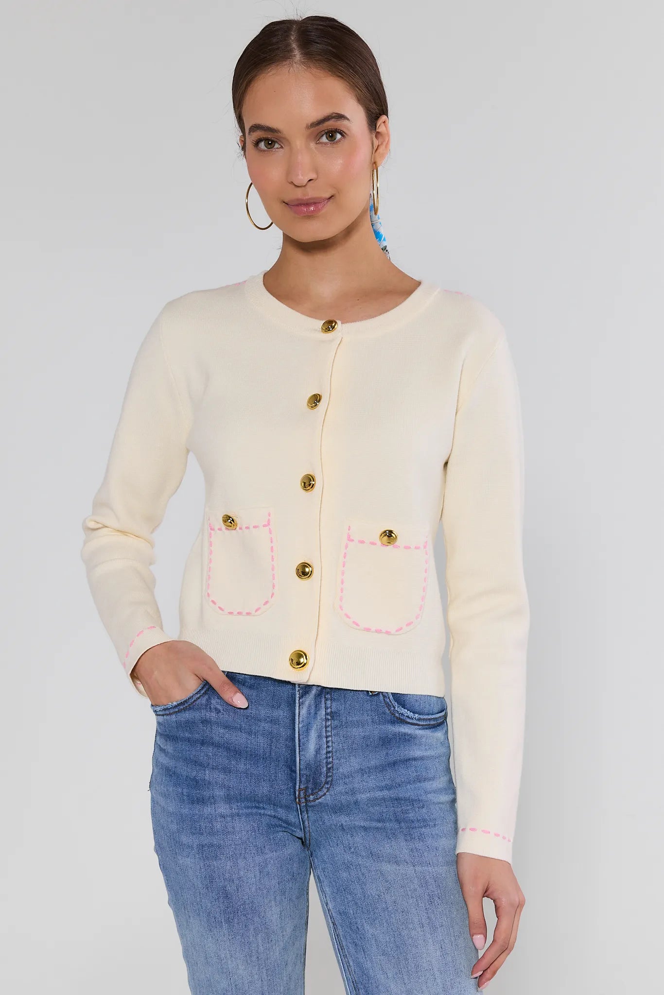 Woman wearing a cream cardigan with gold buttons and blue jeans on a white background