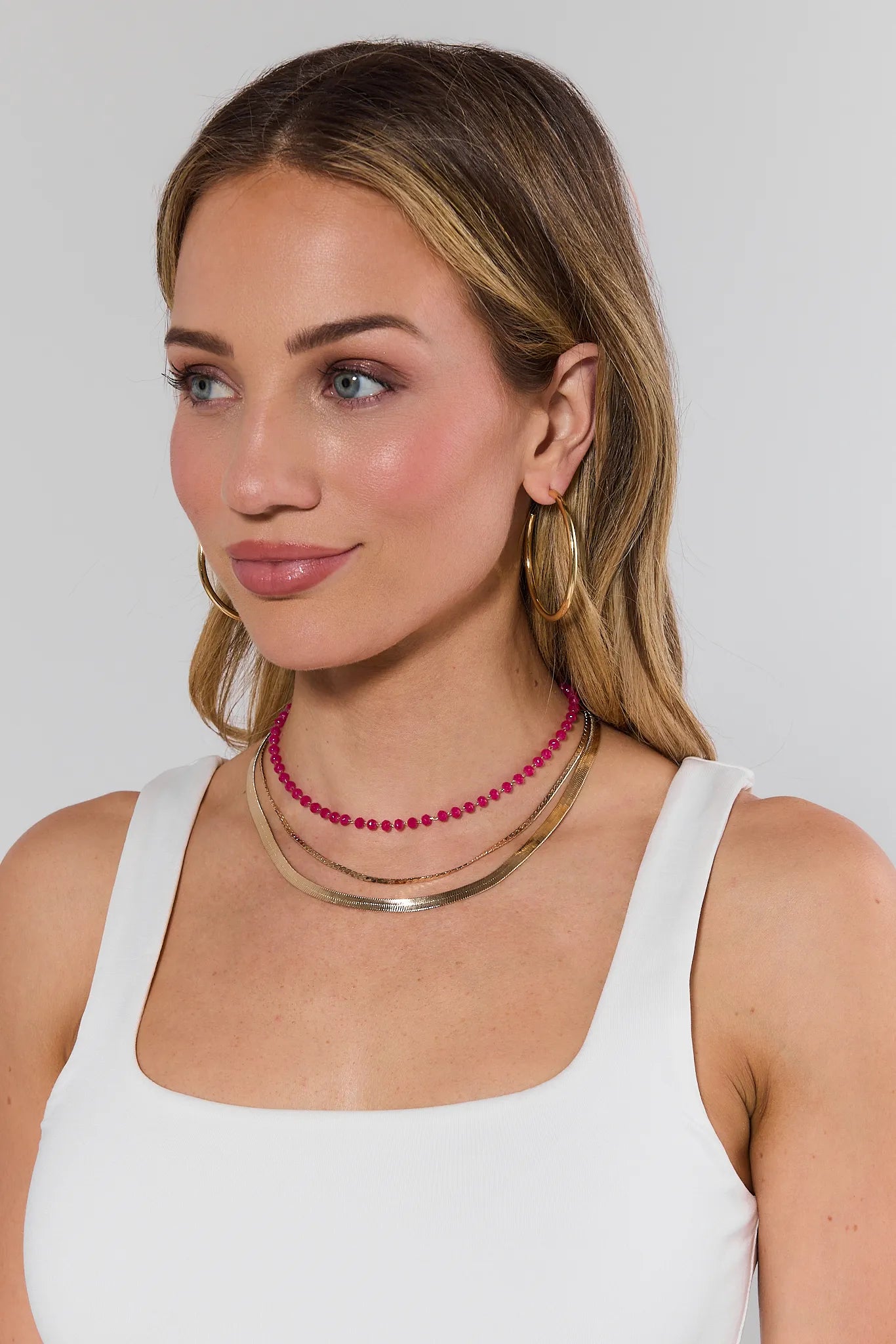 Woman wearing a white tank top and multiple necklaces on a plain background