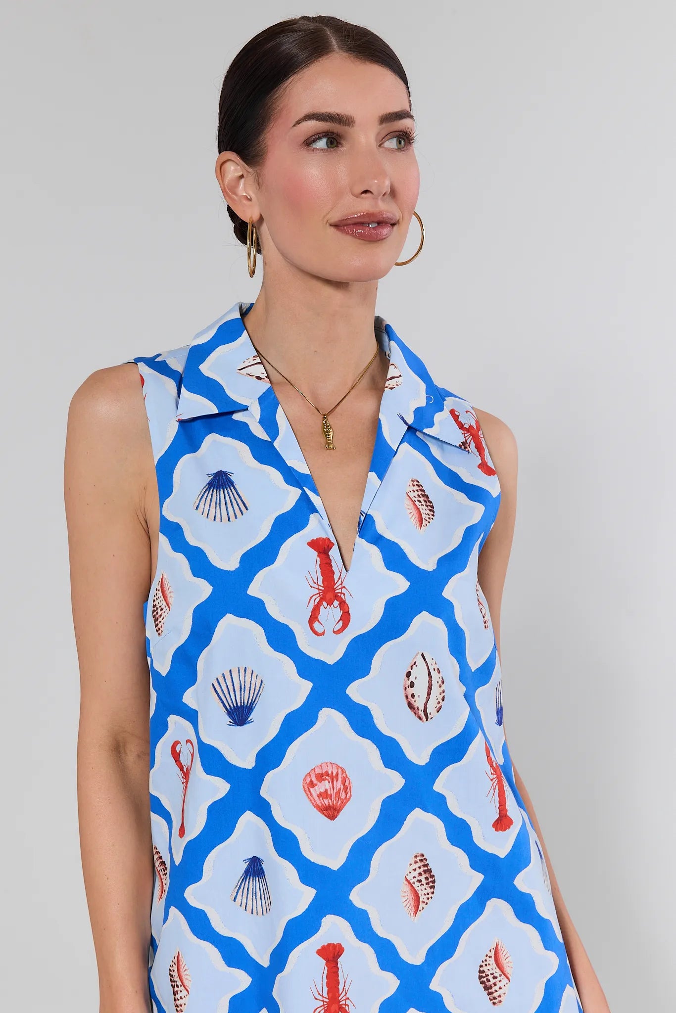 Woman wearing the Ellen Blue Seaside Short Dress with a geometric pattern, featuring lobsters and seashells, accessorized with gold hoop earrings and necklace.