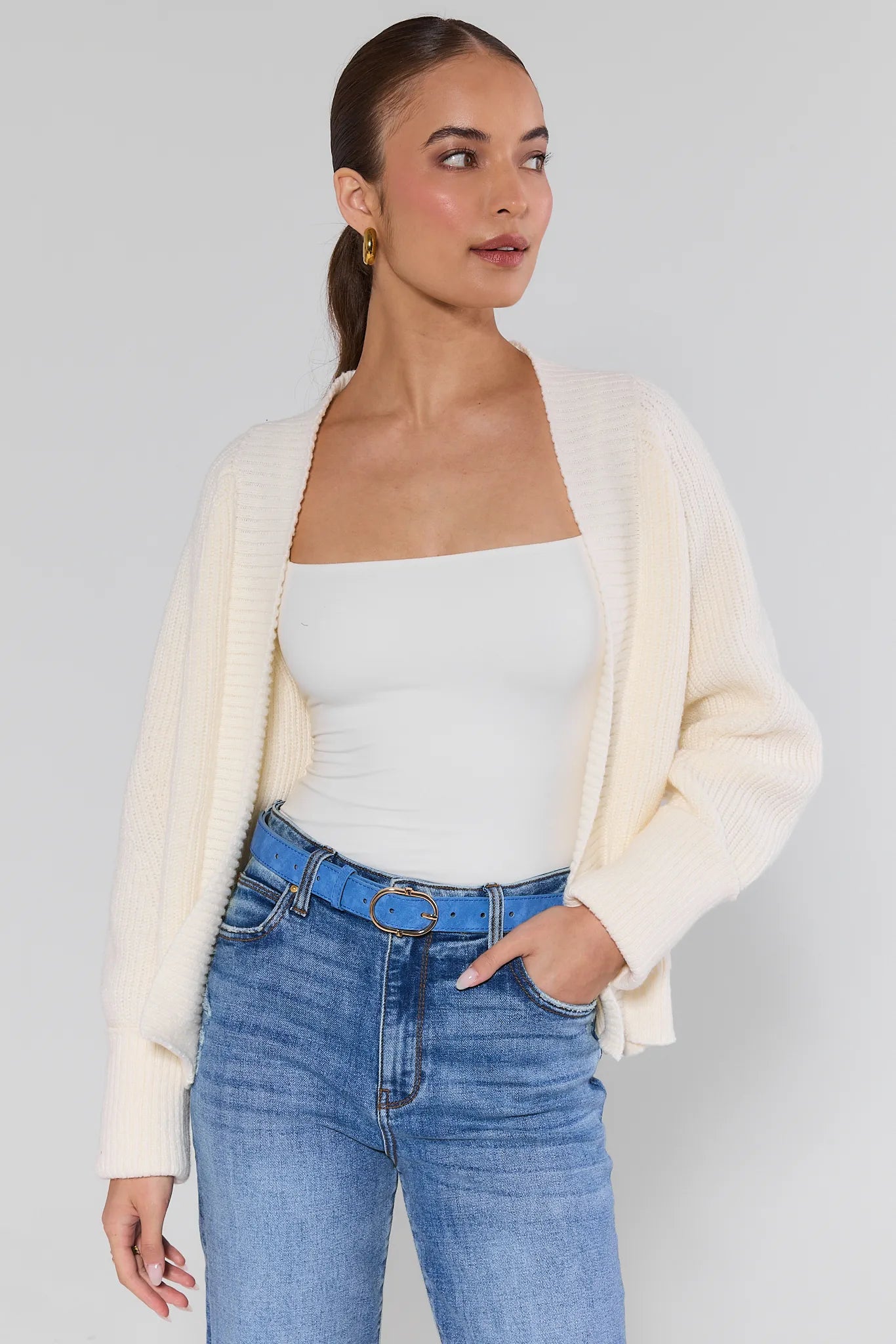 Woman wearing a cream cardigan, white top, Trisha Blue Faux Suede Belt, and blue jeans on a plain background