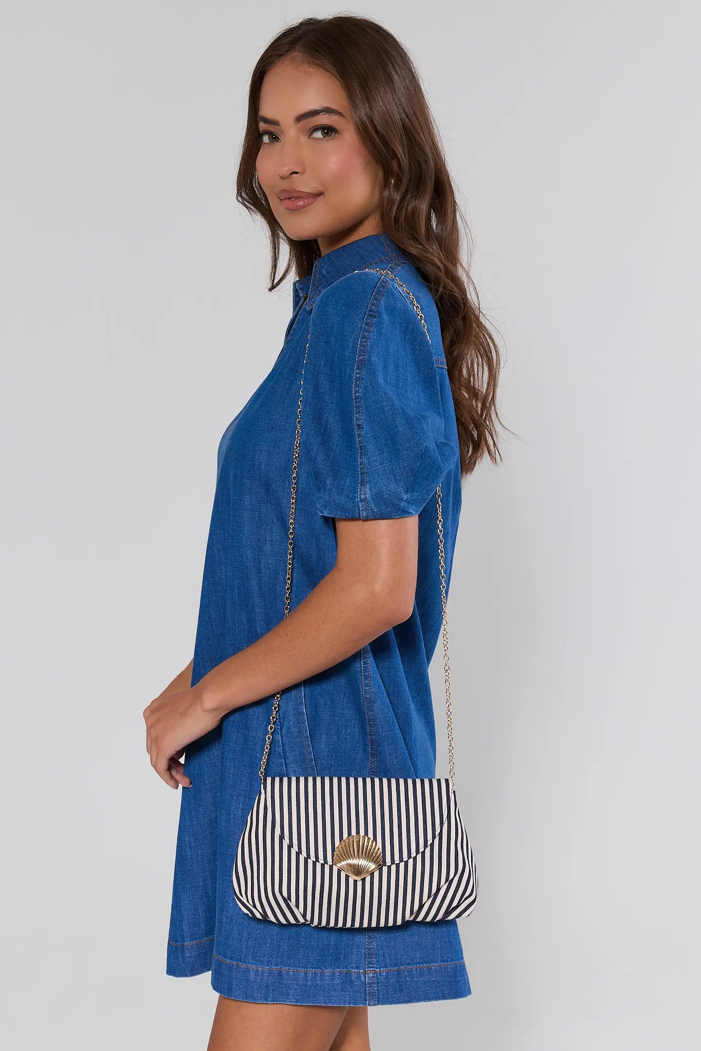Woman wearing a blue denim dress holding a striped handbag against a plain background