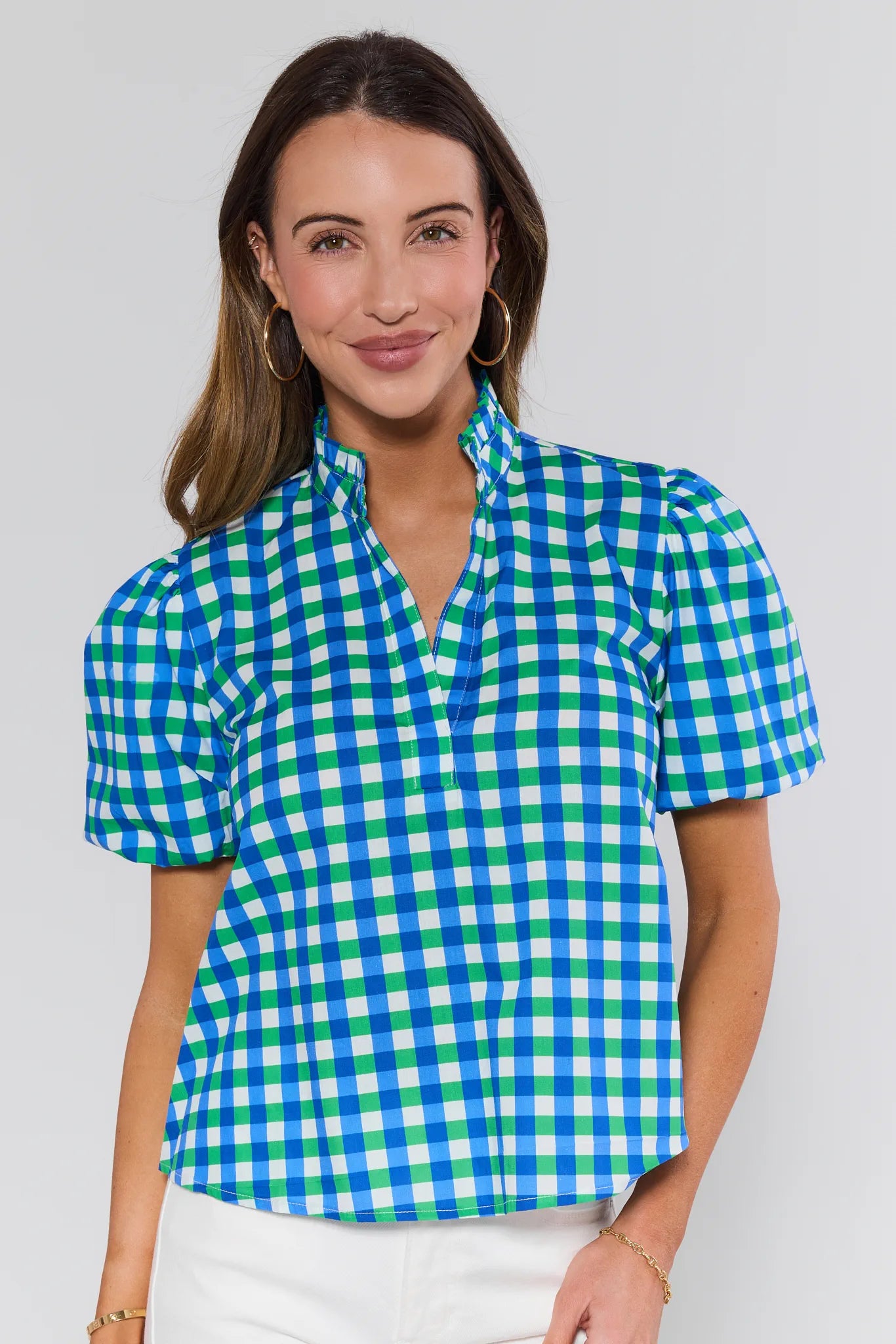 Young woman in a Kathleen Mixed Gingham Short Sleeve Top with puff sleeves, paired with white pants, gold hoop earrings, and a bracelet, smiling gently.