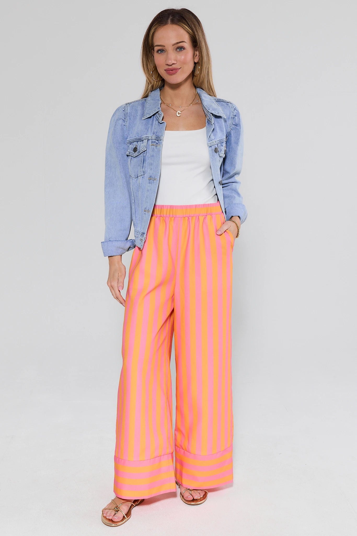 Woman wearing a light blue denim jacket, white top, and pink striped pants on a white background