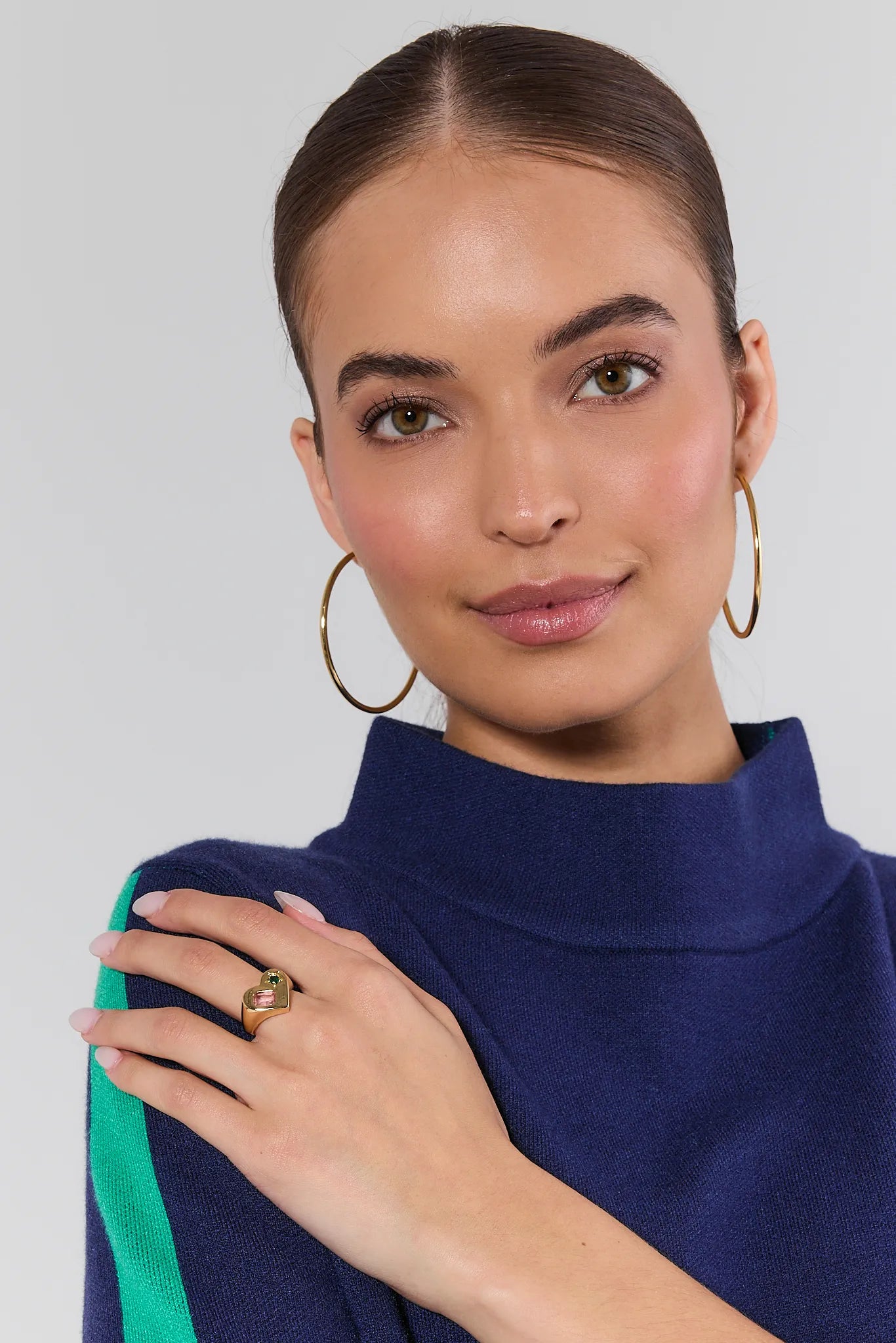 Woman wearing gold hoop earrings and a Renee Gold Multi Stone Heart Ring, holding a blue garment against a gray background