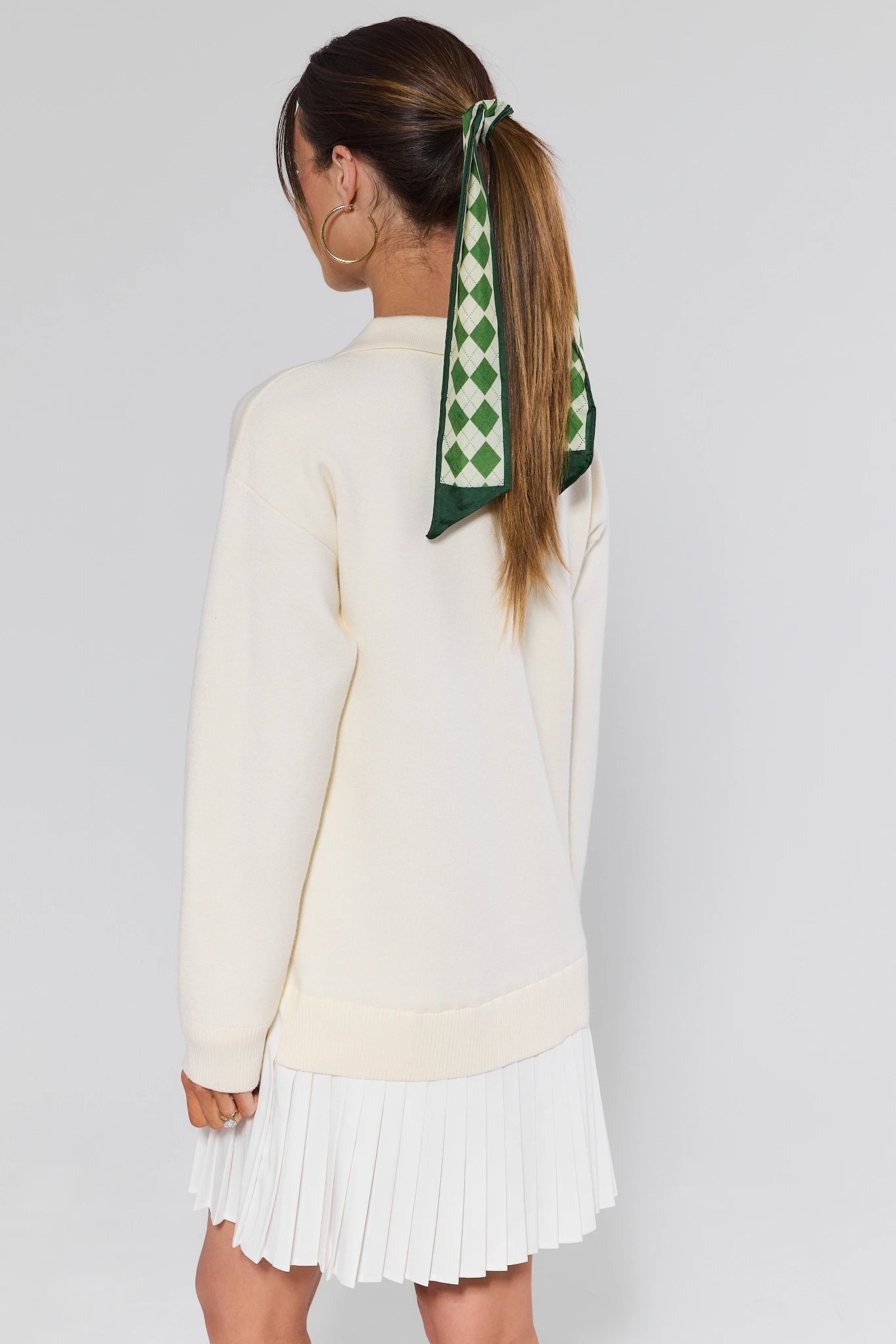 Woman wearing a cream sweater and white pleated skirt with a green and white checkered scarf on her head against a light gray background.