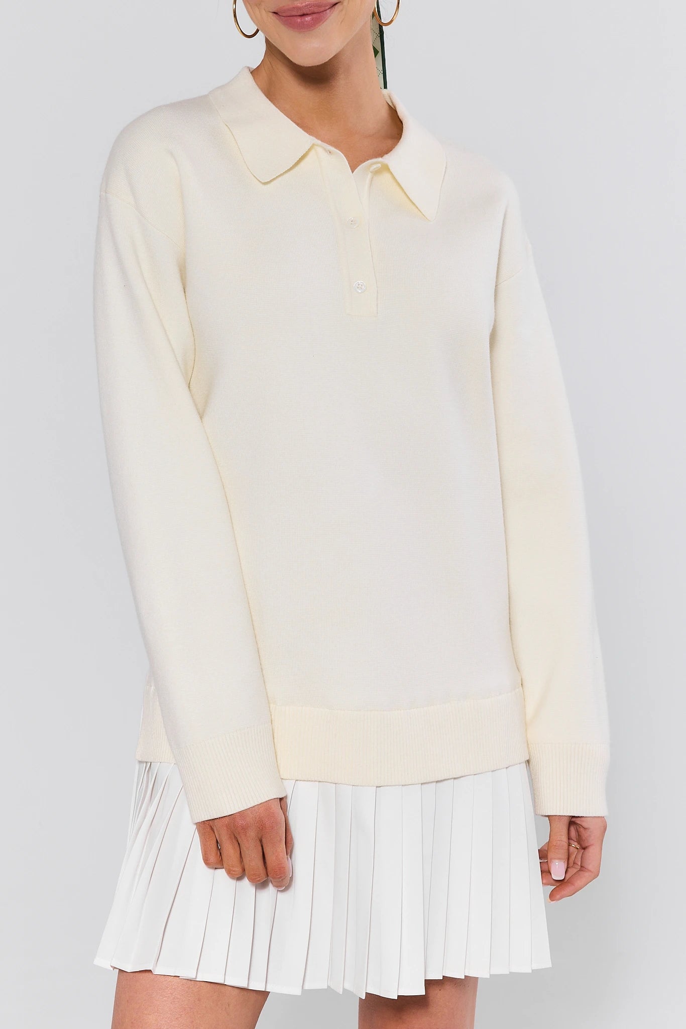 Person wearing a light-colored sweater and white pleated skirt on a plain background
