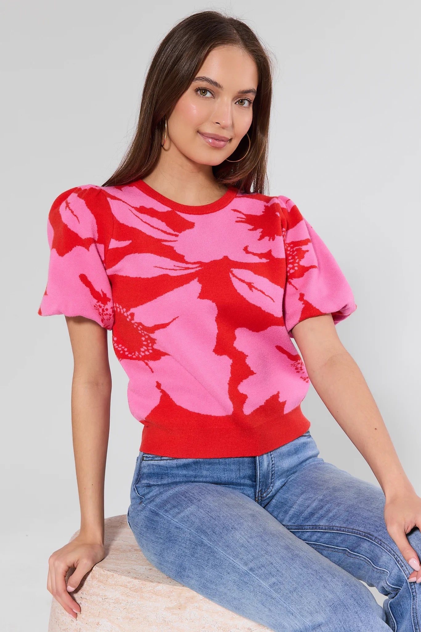 woman wearing pink and red floral short sleeve top sitting on a stool with a white background