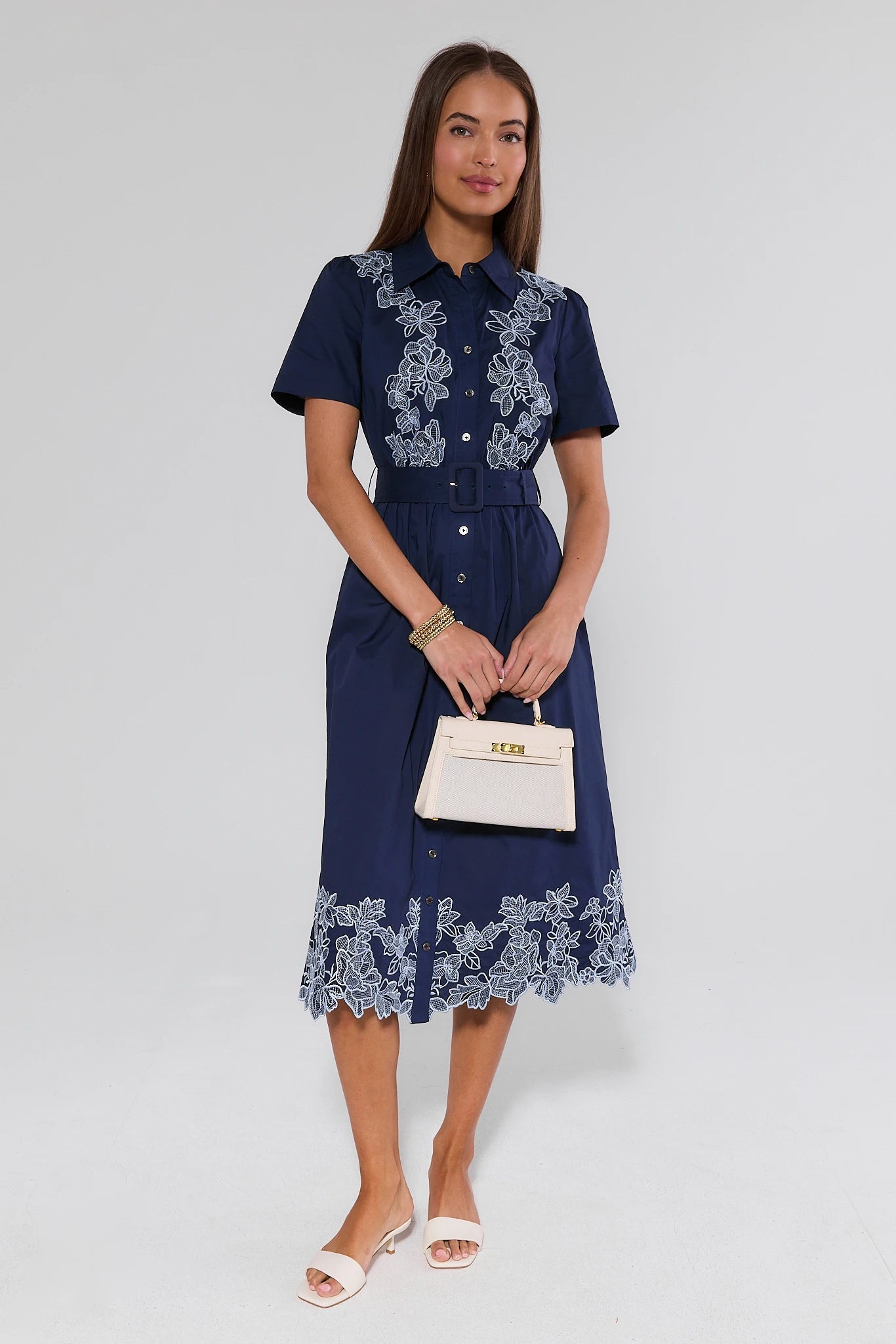 Woman wearing a navy dress with white lace details, holding a white handbag on a light gray background
