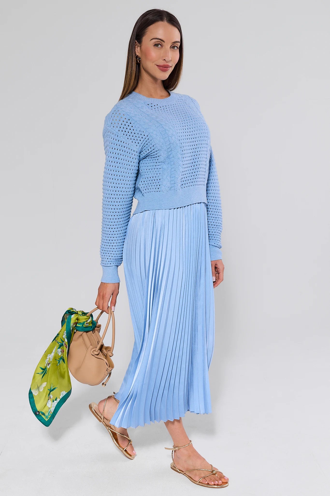 Woman wearing a light blue sweater and pleated skirt holding a beige handbag with a green scarf.