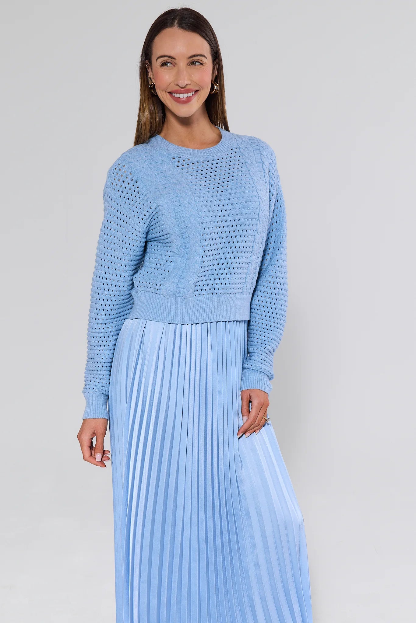 Woman wearing a light blue knitted sweater and pleated skirt against a plain background