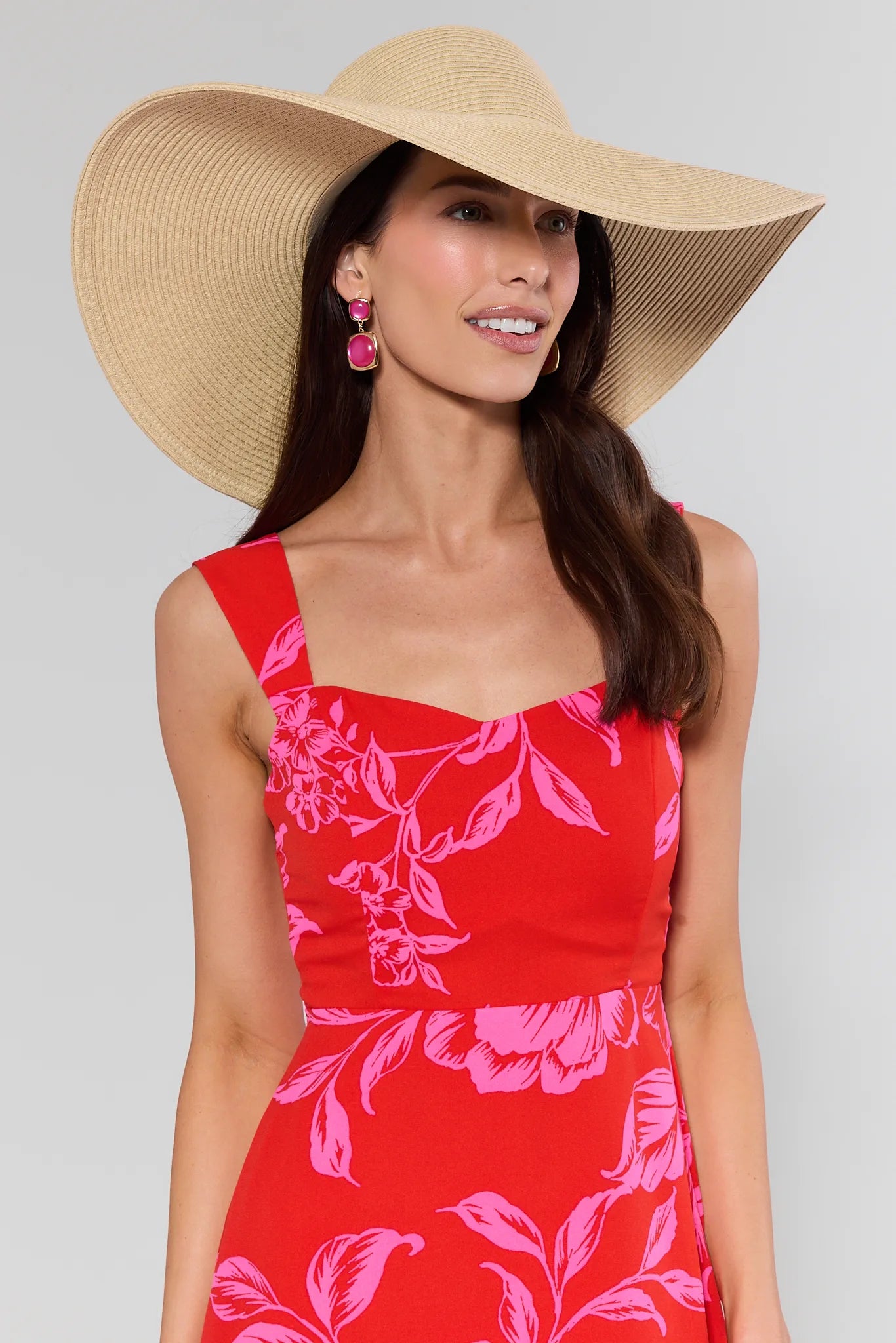 Woman wearing the Betty Raffia Wide Brim Sun Hat, featuring a woven straw finish and floppy brim, paired with a sleeveless floral dress and gem-tone earrings.