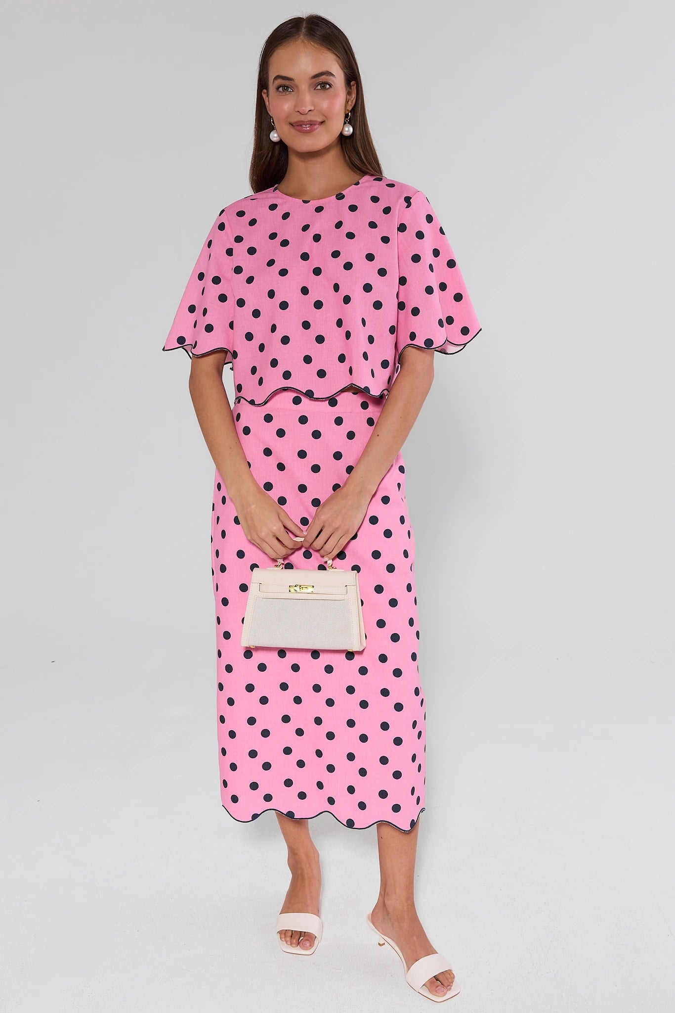 Woman wearing a pink polka dot dress holding a beige clutch against a gray background