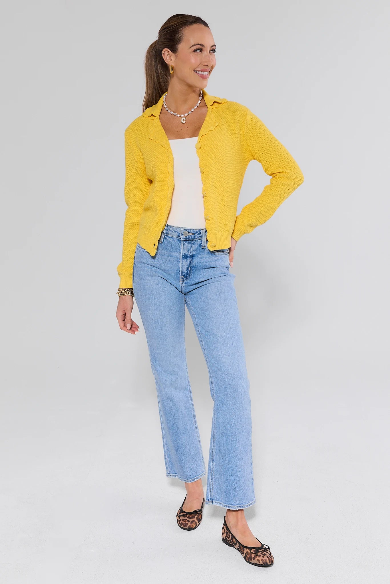 Woman wearing a yellow cardigan, white top, and blue jeans on a light gray background