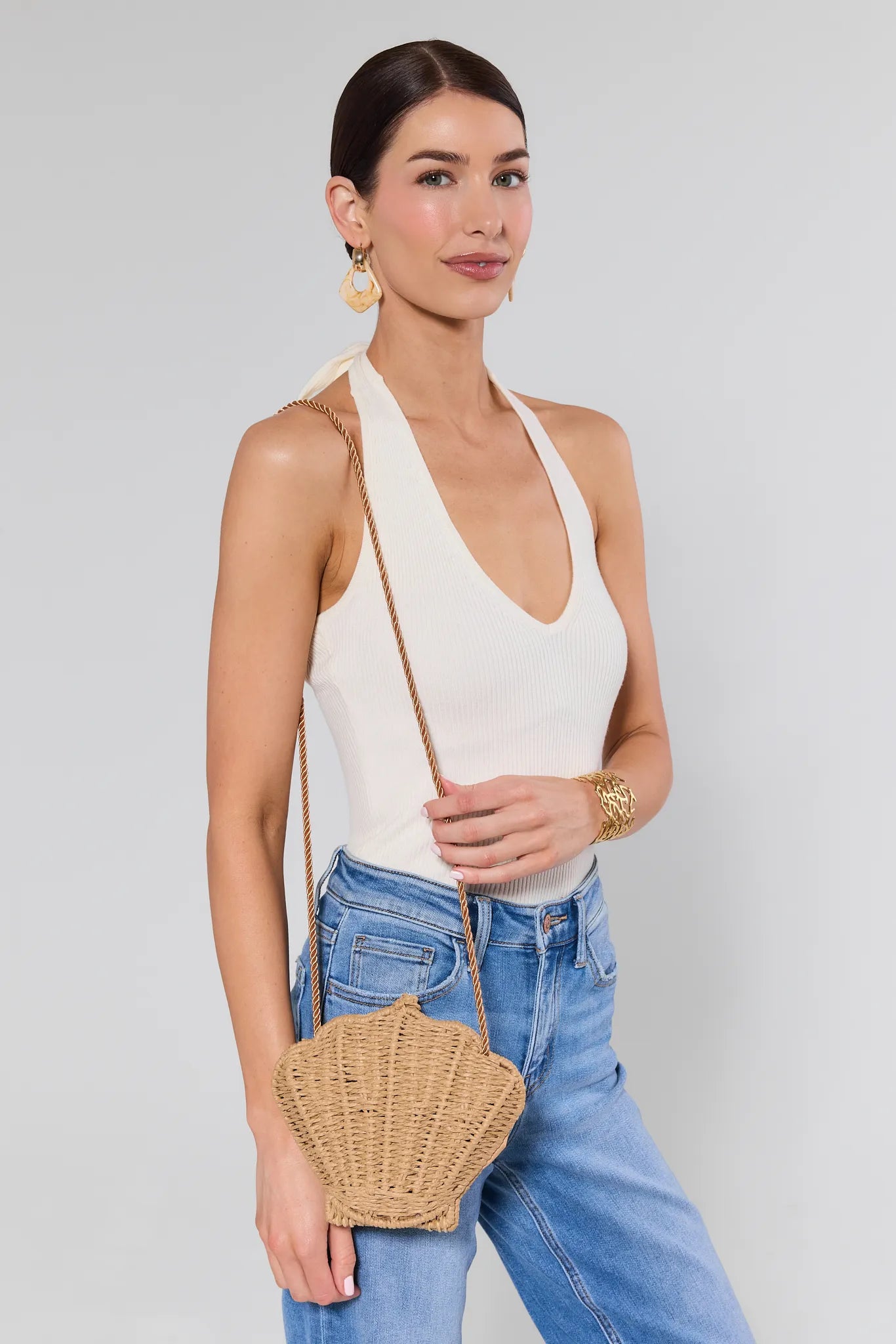 Woman wearing a white halter top and blue jeans, holding a straw bag against a plain background