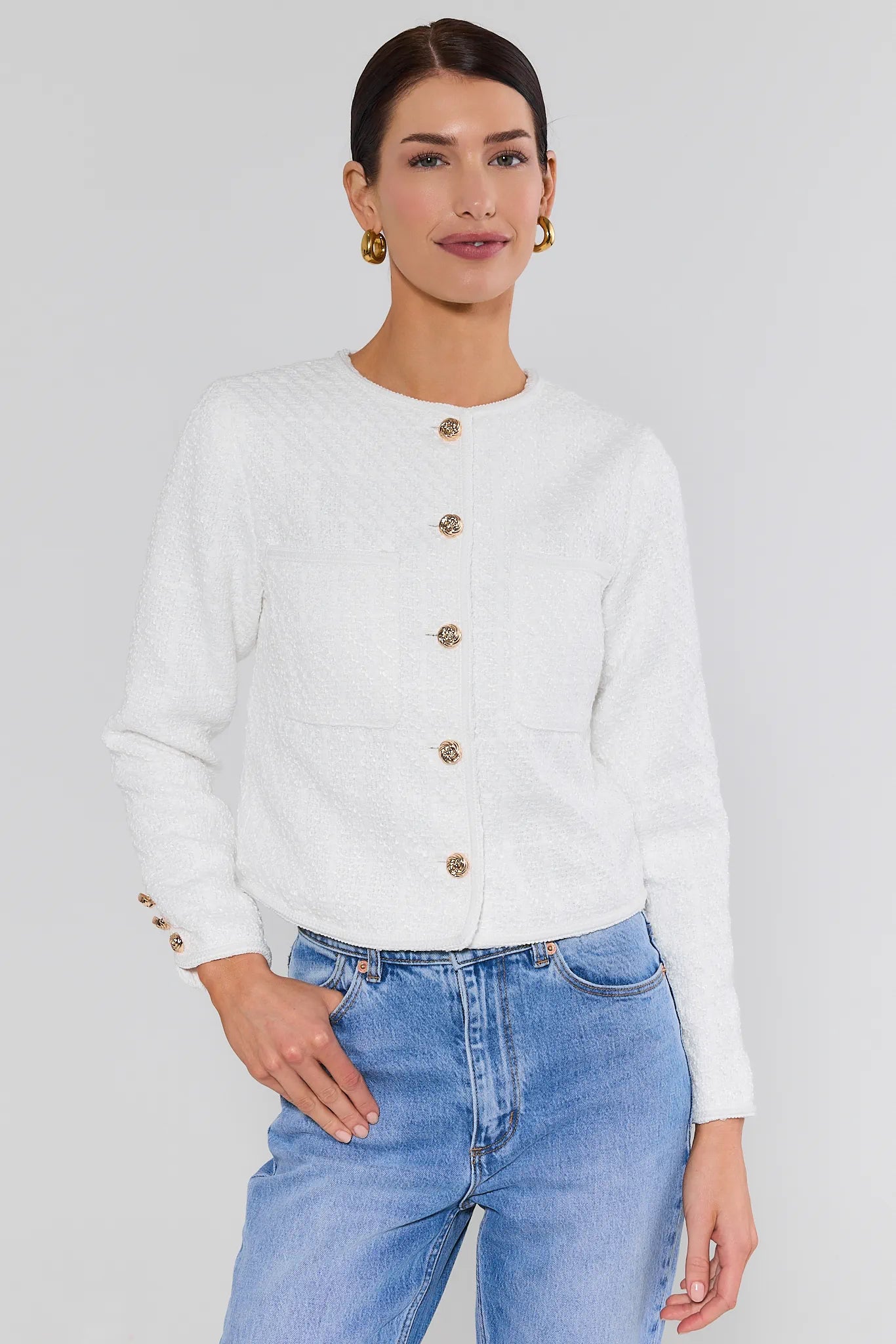 Woman wearing a white cardigan with gold buttons and blue jeans on a plain background