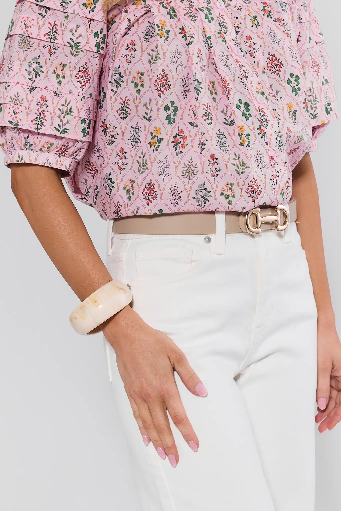 Person wearing a floral blouse and white pants with a beige belt, on a plain background