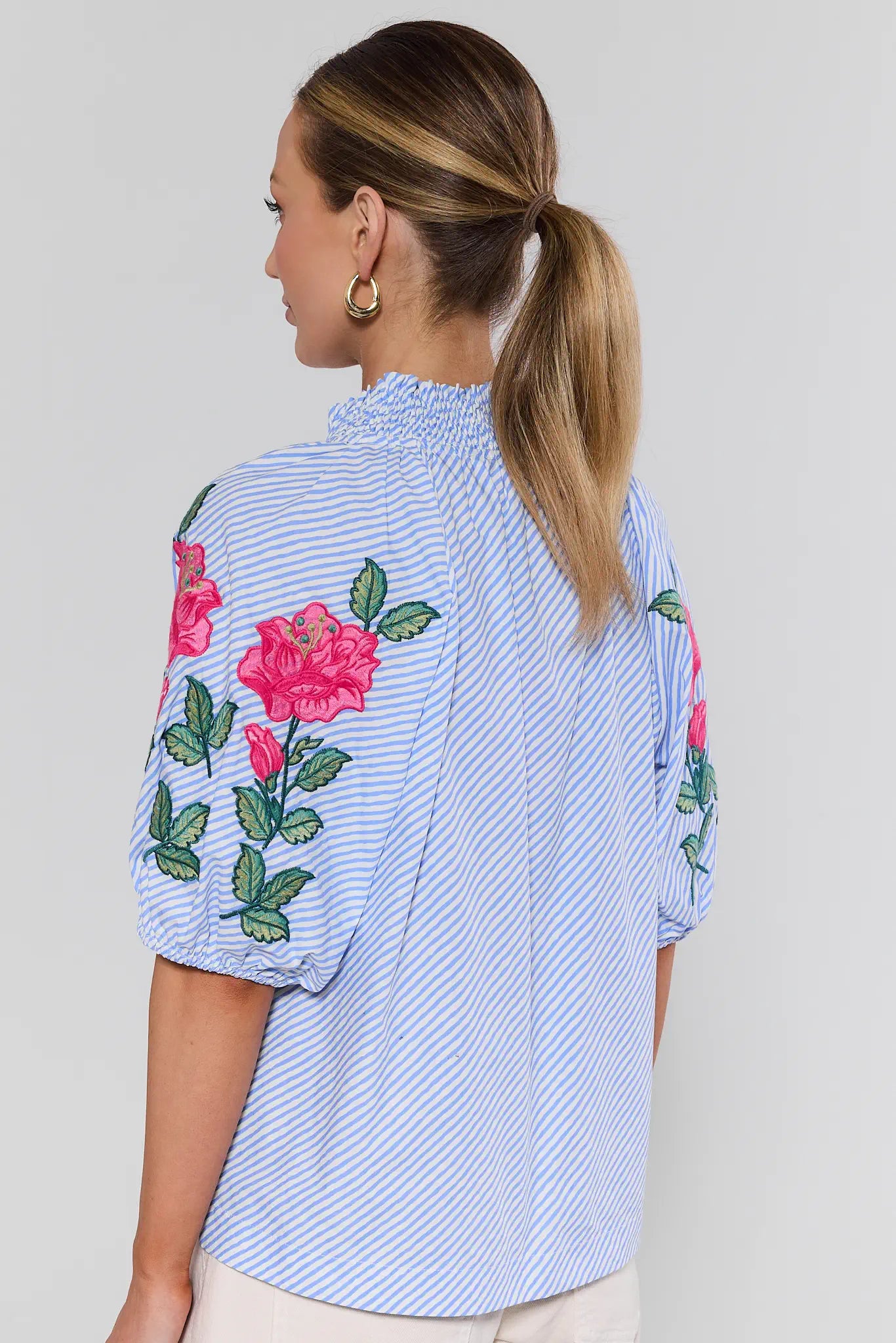 Woman wearing a blue striped shirt with floral embroidery on the back, standing against a plain background.
