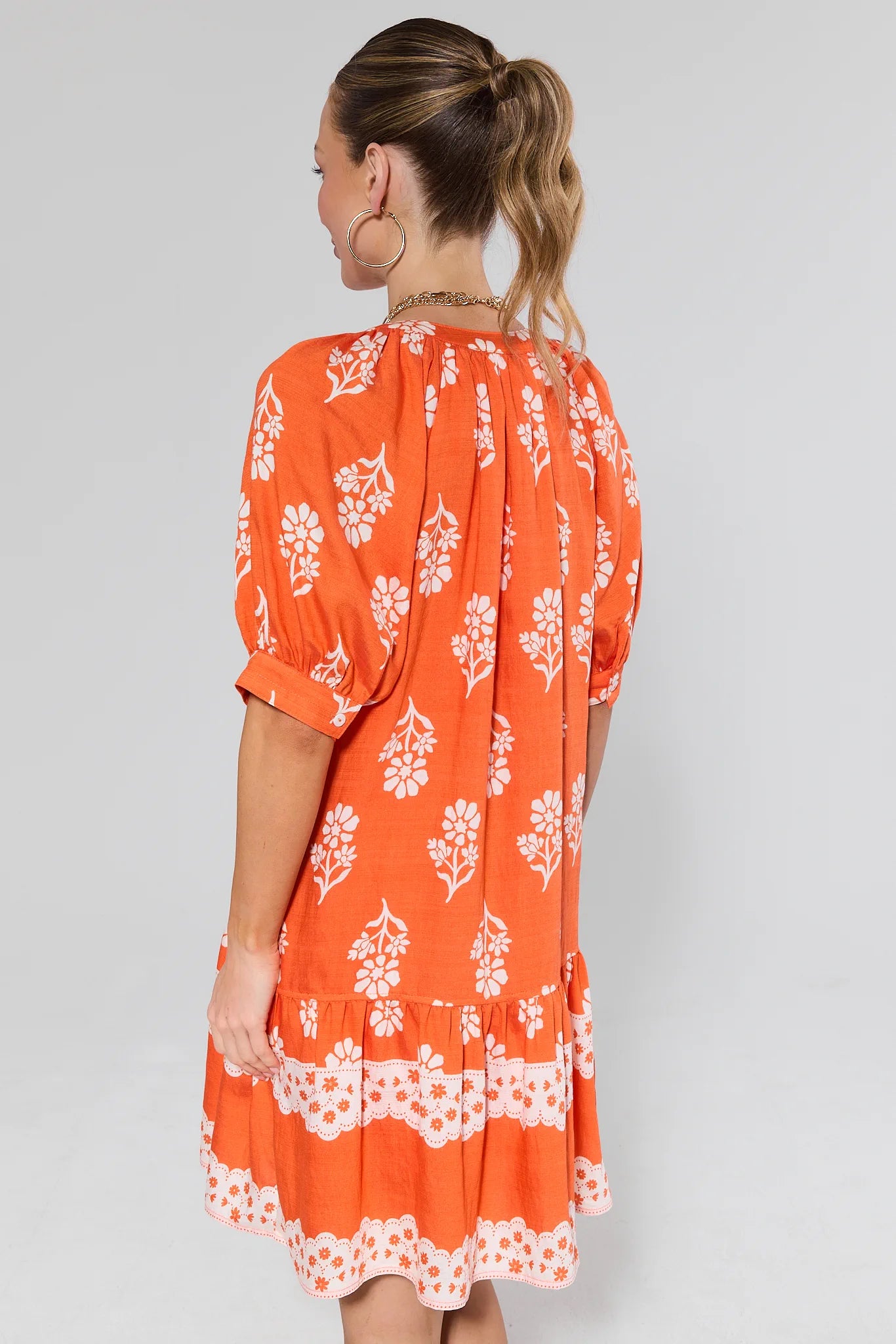 Woman standing wearing an orange dress with white floral pattern on a plain background