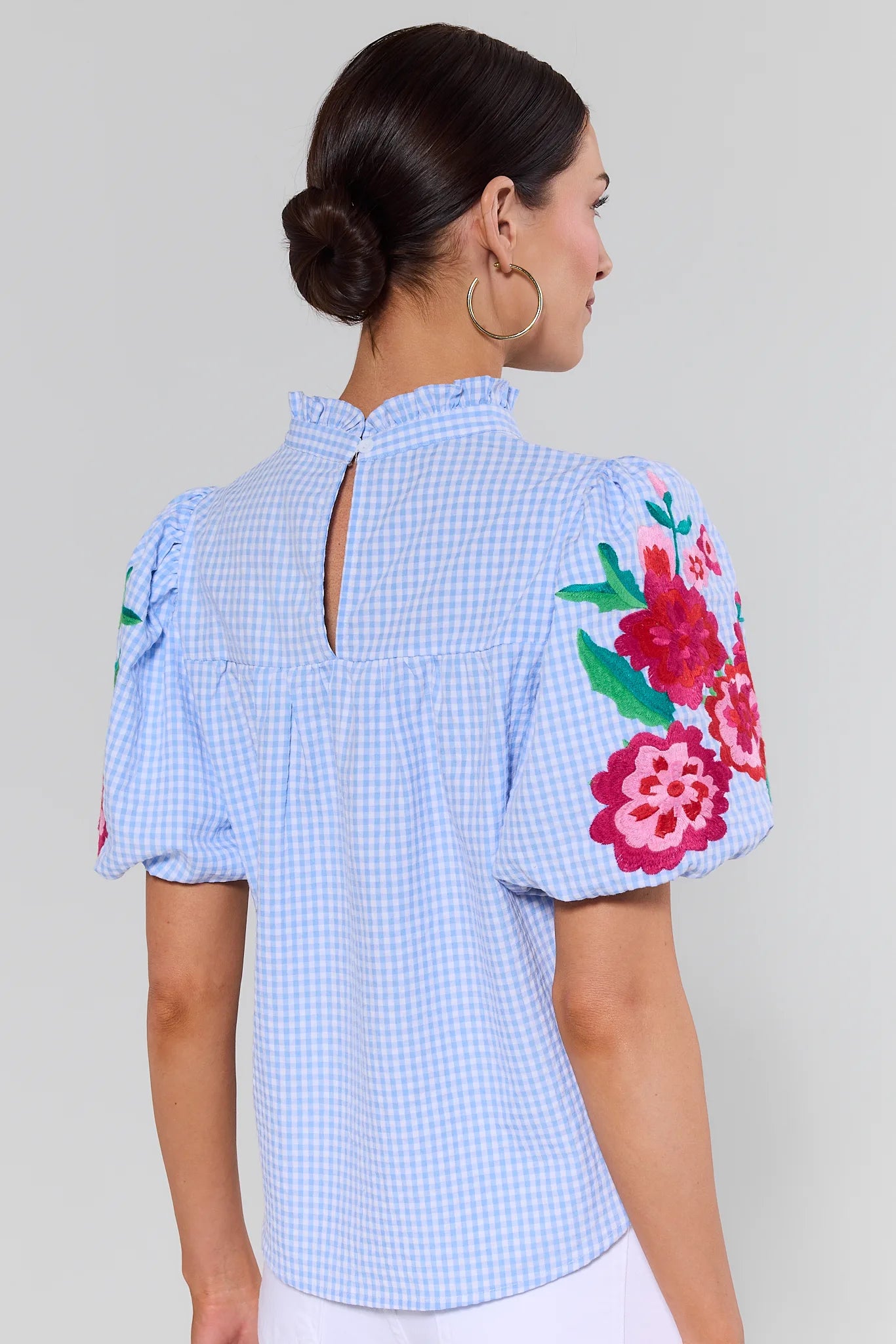 Woman standing wearing a blue checkered blouse with floral embroidery on a plain background