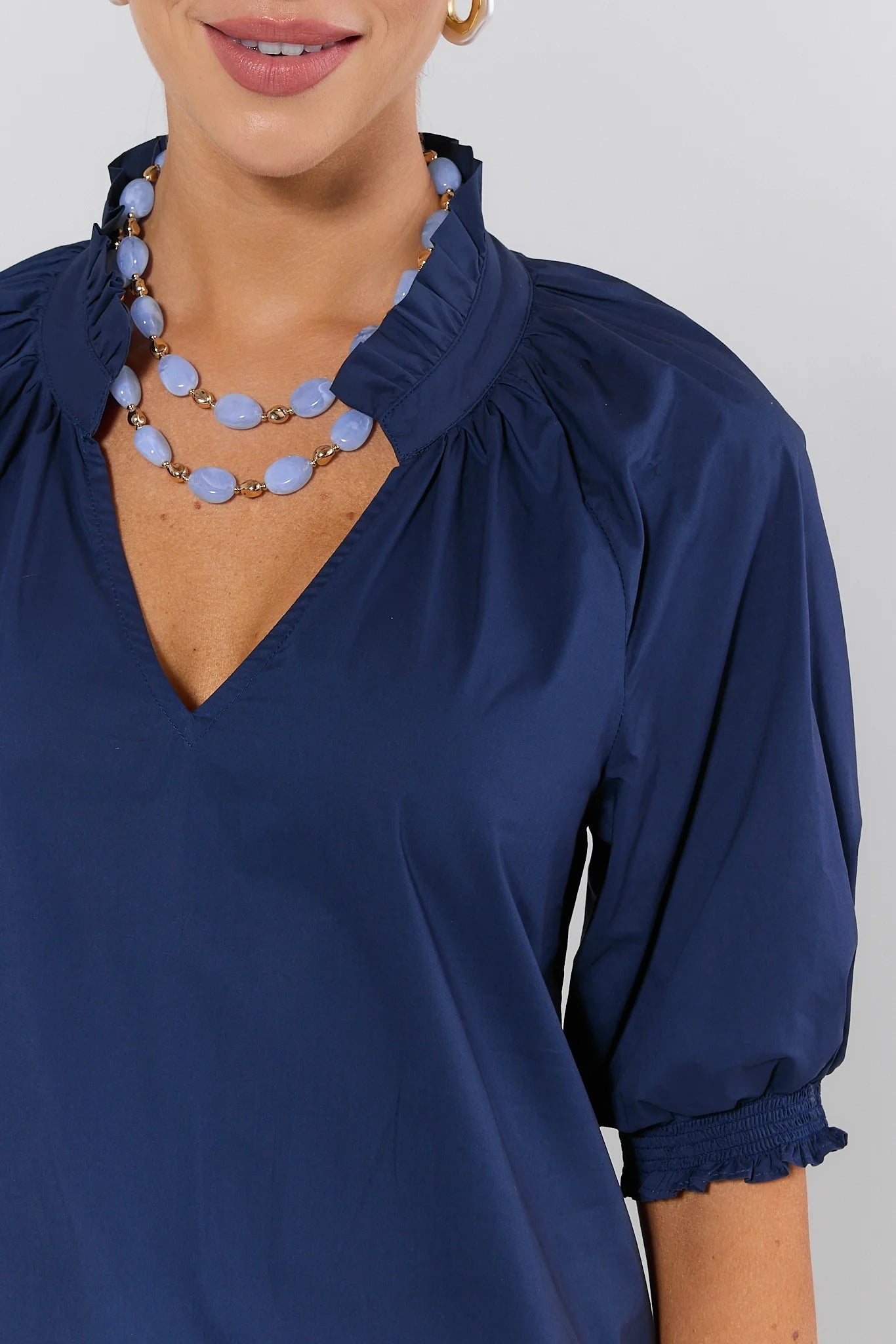 Woman wearing a blue blouse with ruffled collar and cuffs, and a necklace on a plain background