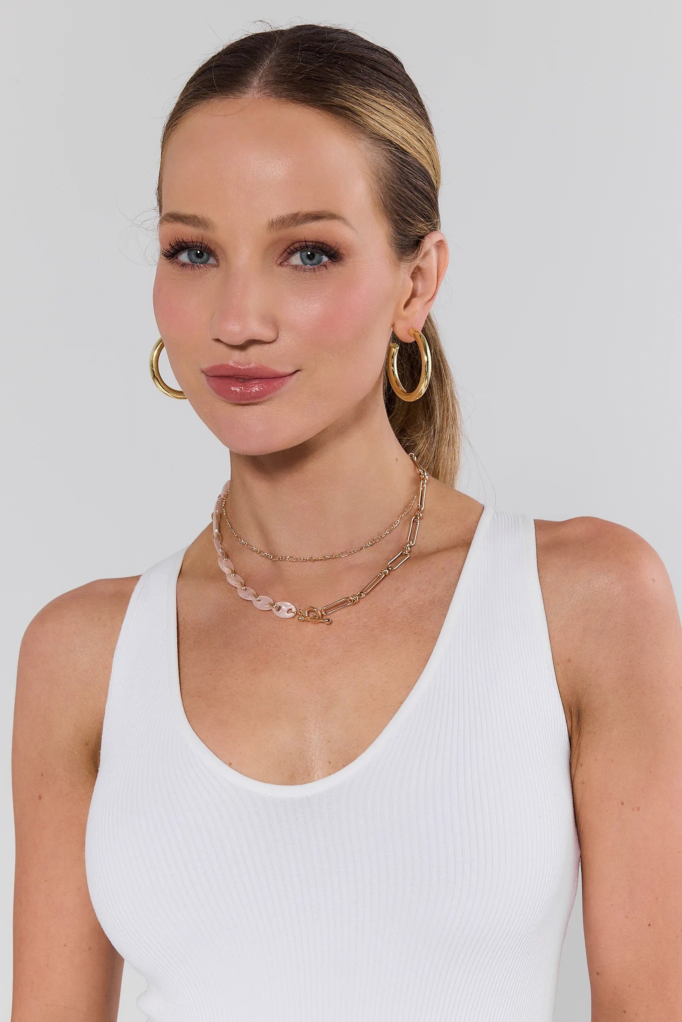 Woman wearing gold hoop earrings and layered necklaces against a white background