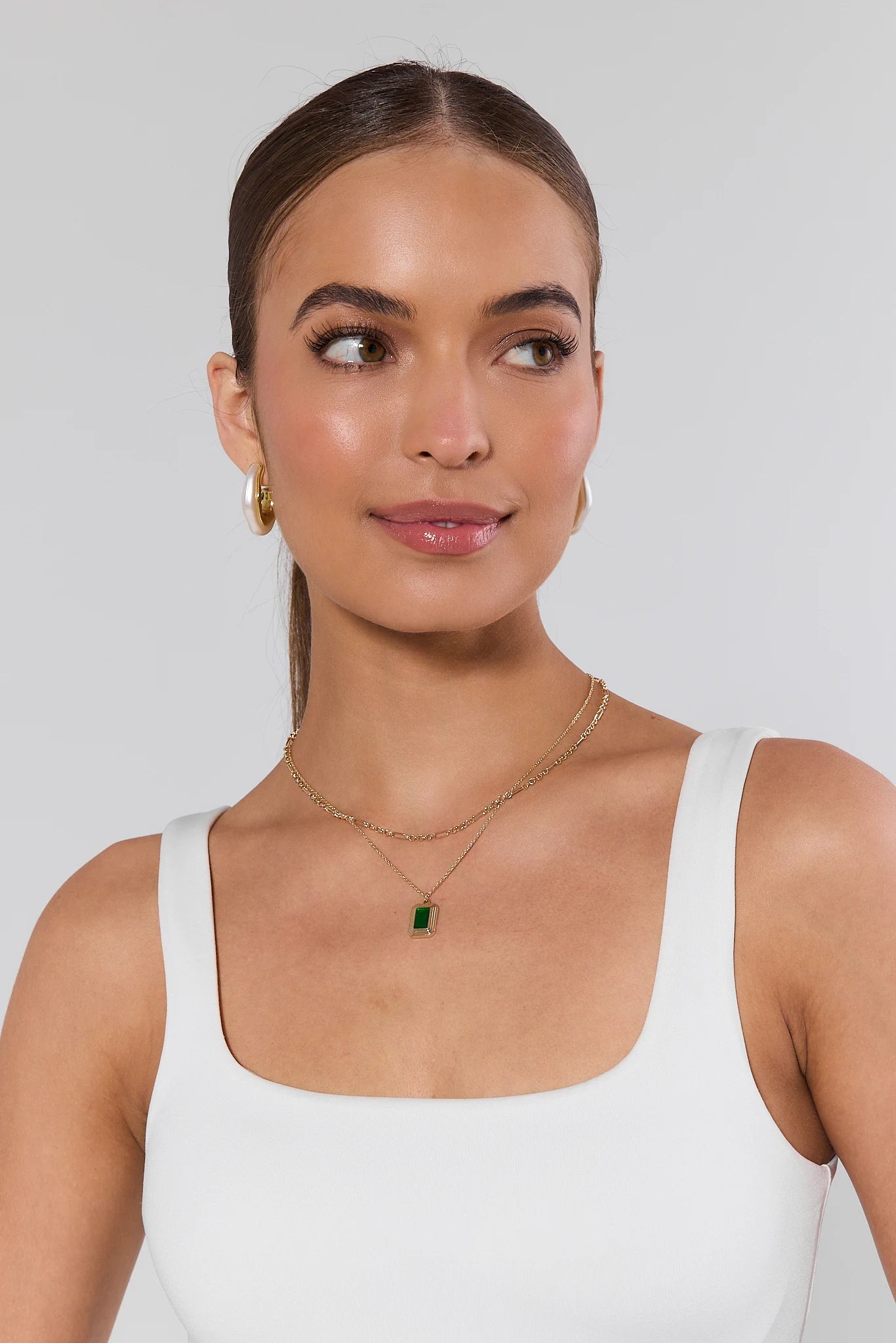 Woman wearing a white tank top and gold necklace with a green pendant against a light gray background