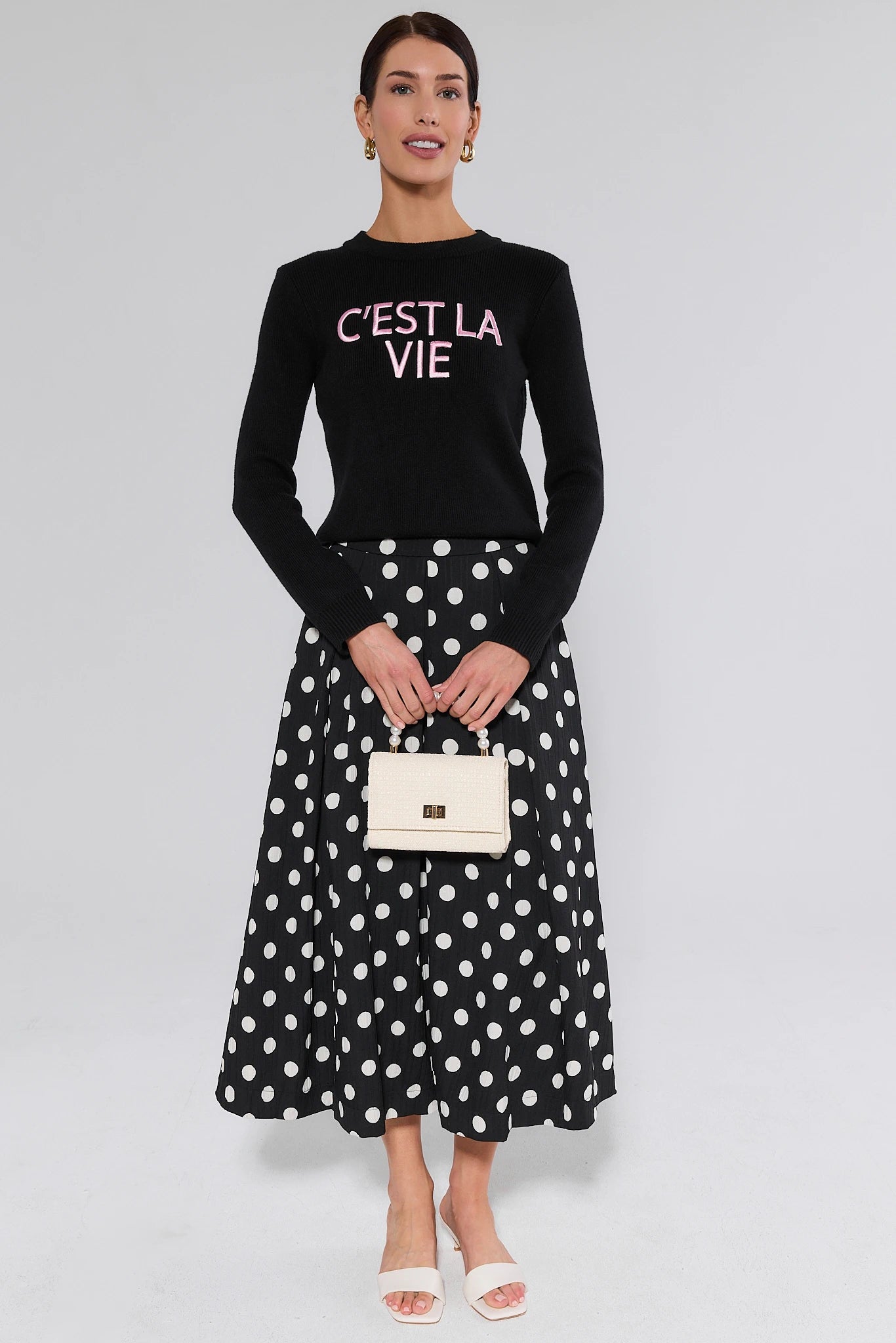 Woman wearing a black sweater with 'C'EST LA VIE' text and a black polka dot skirt, holding a beige handbag on a gray background.