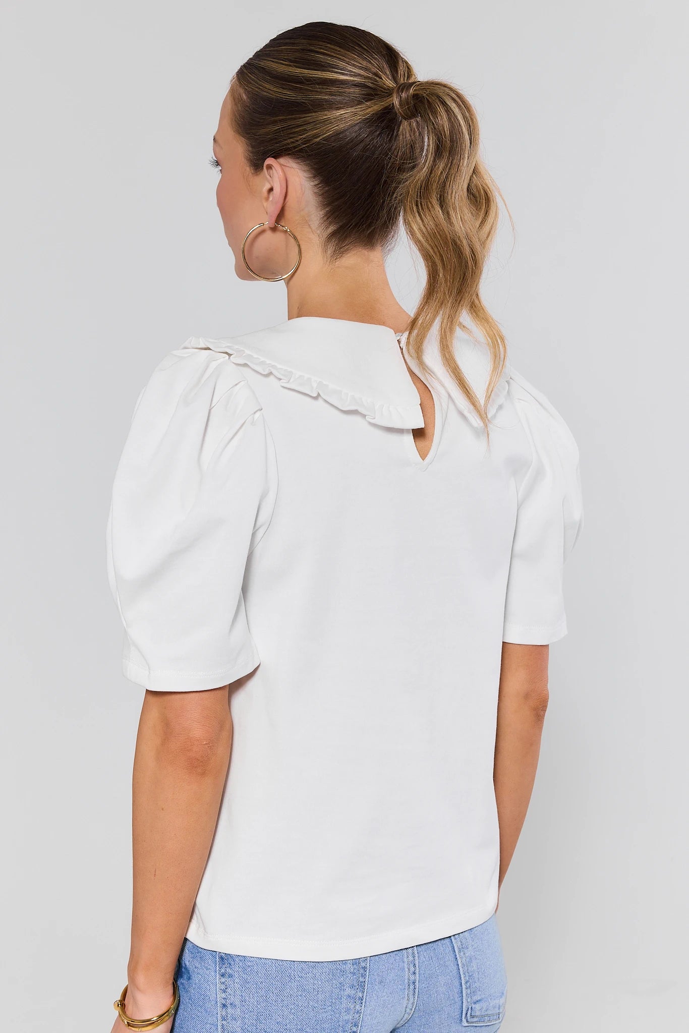 Woman standing wearing a white blouse with ruffled shoulders against a plain background