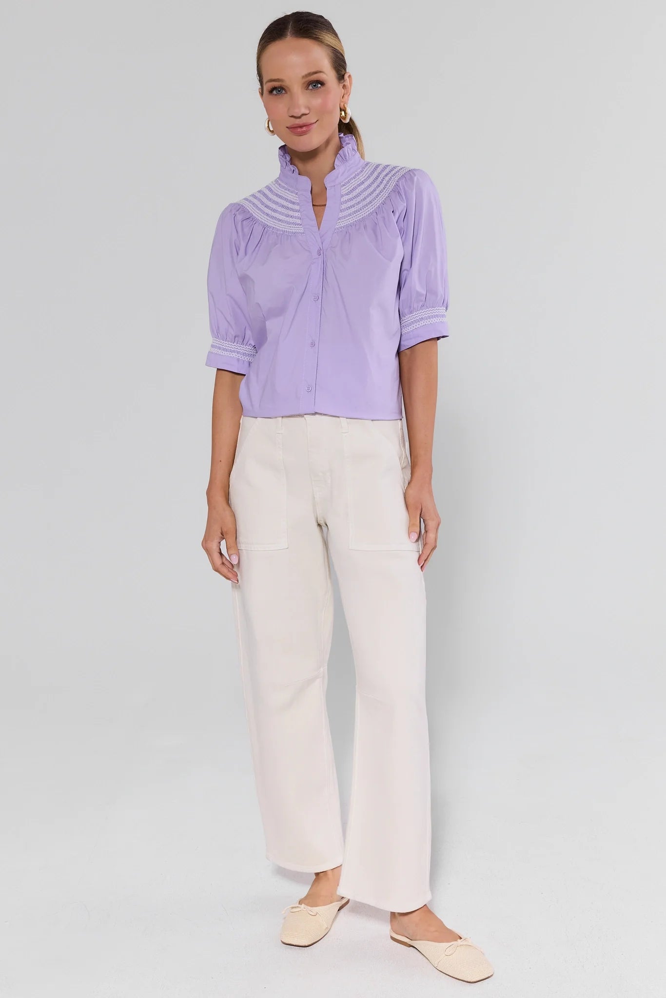 Woman wearing a light purple blouse with a collar and white pants on a plain background