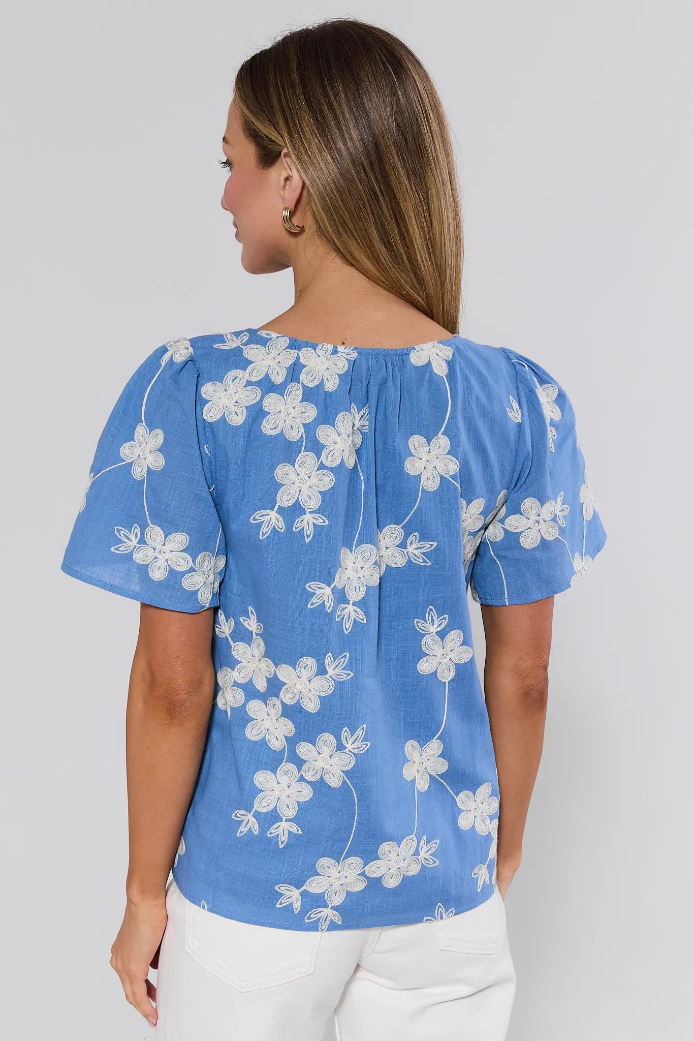 Back view of the Cindy Blue Embroidered Short Sleeve Top, showcasing white floral embroidery, short sleeves, and relaxed fit, paired with white pants.