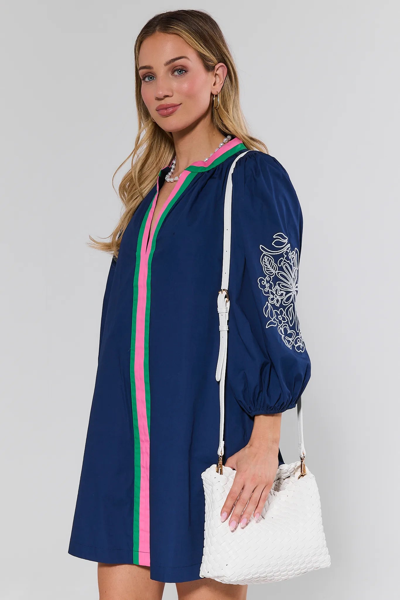 Woman wearing a navy blue dress with pink and green accents, holding a white handbag on a light gray background