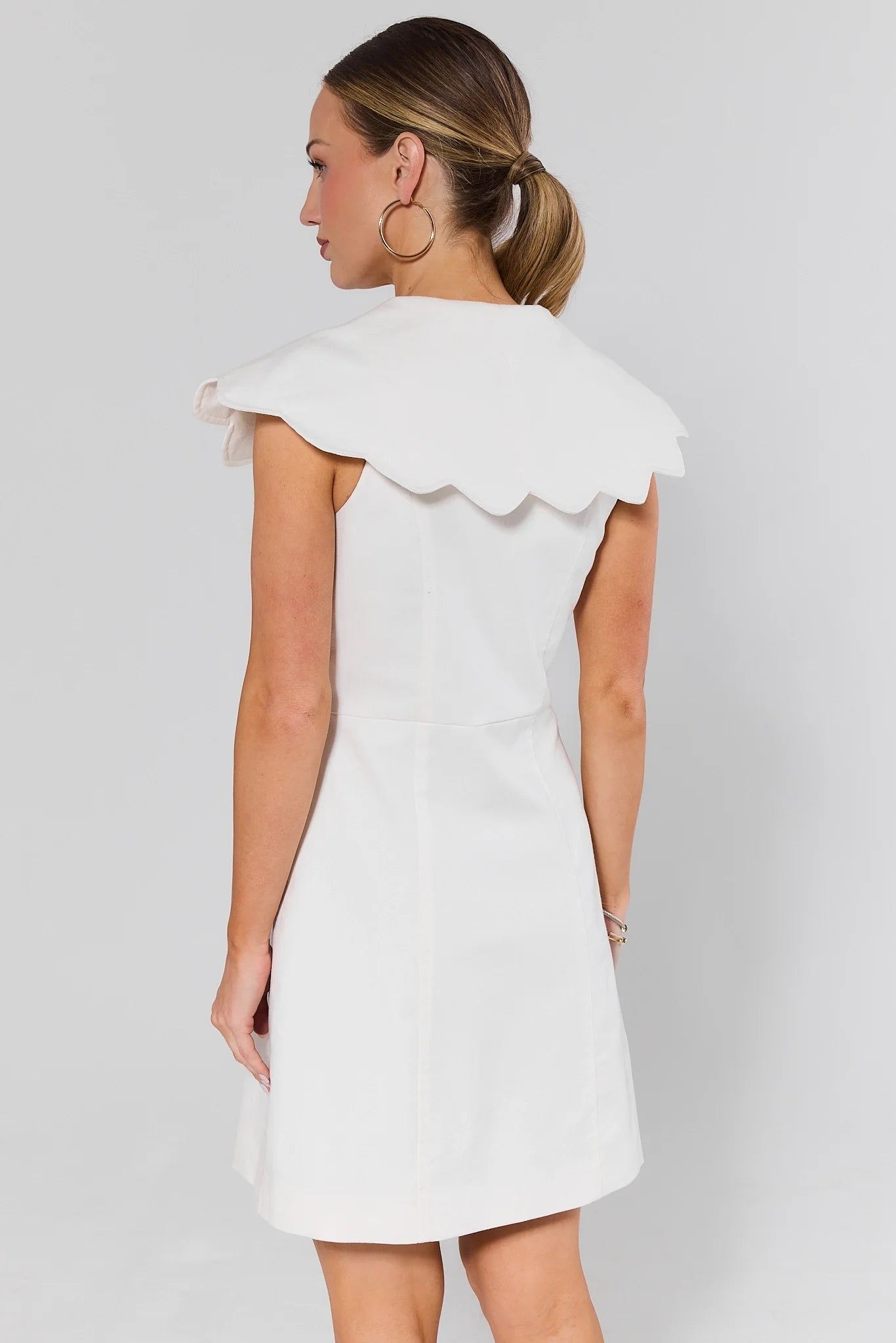 Woman standing wearing a white dress with a scalloped collar against a plain background