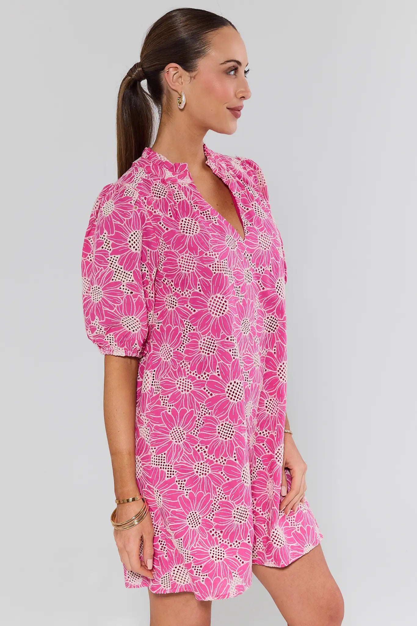 Woman modeling Bianca Pink Floral Eyelet Short Dress with daisy motif, puff sleeves, V-neck, and mid-thigh length, accessorized with gold hoop earrings and bangles.