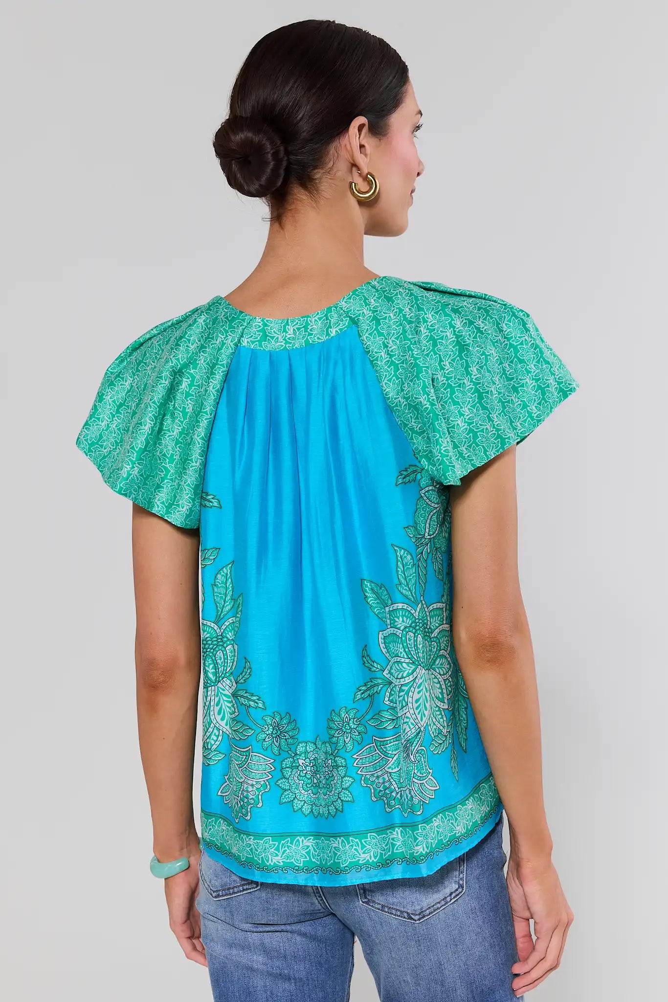 Woman standing wearing a turquoise blouse with green patterns on a plain background