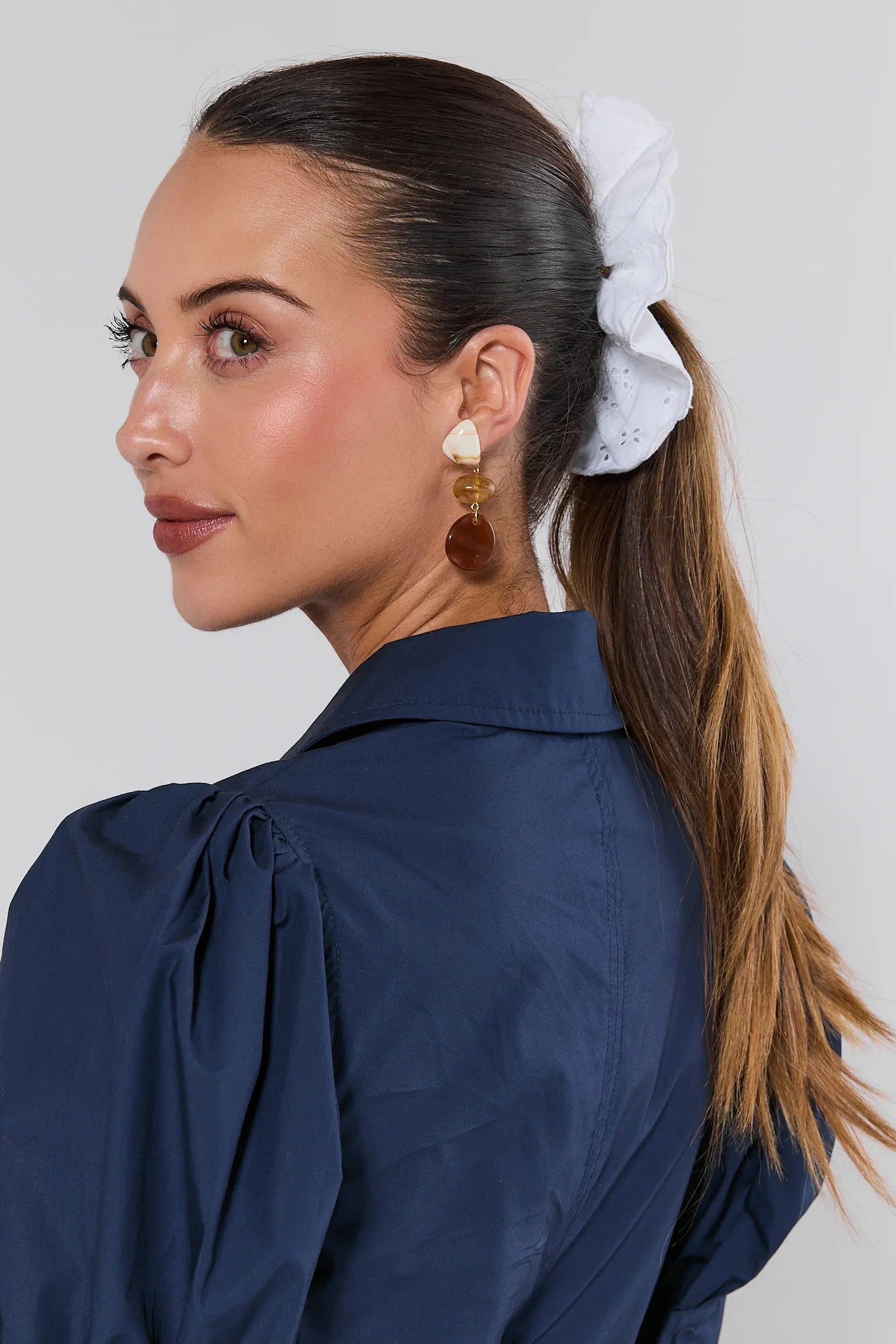 Woman with a ponytail wearing a white scrunchie, blue shirt, and gold earrings on a light gray background