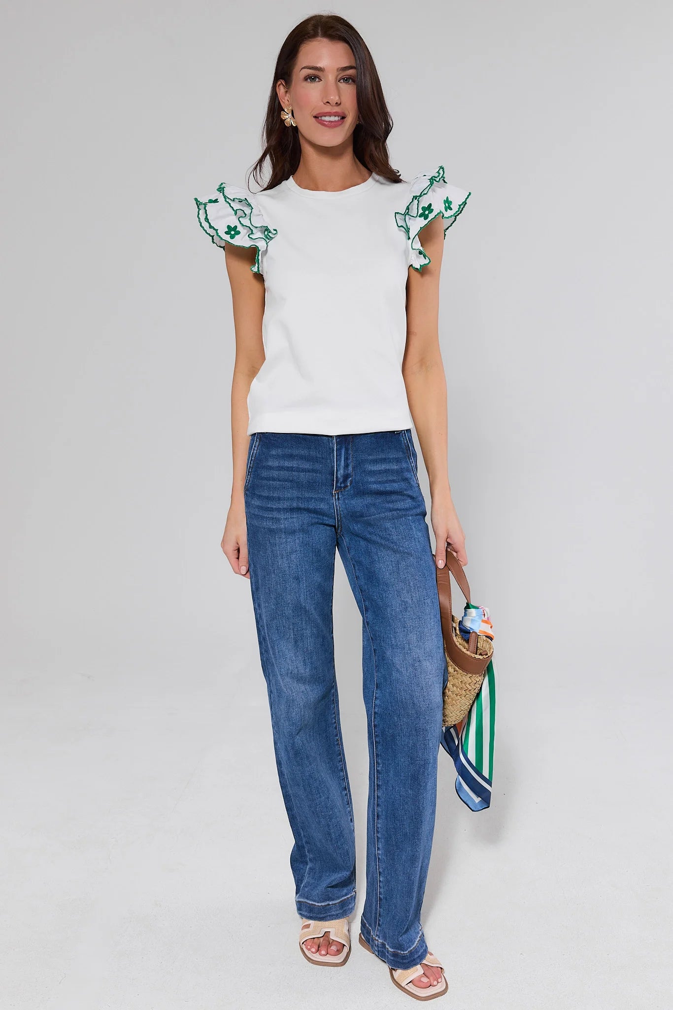 Woman wearing a white top with green floral sleeves and blue jeans, holding a straw bag.
