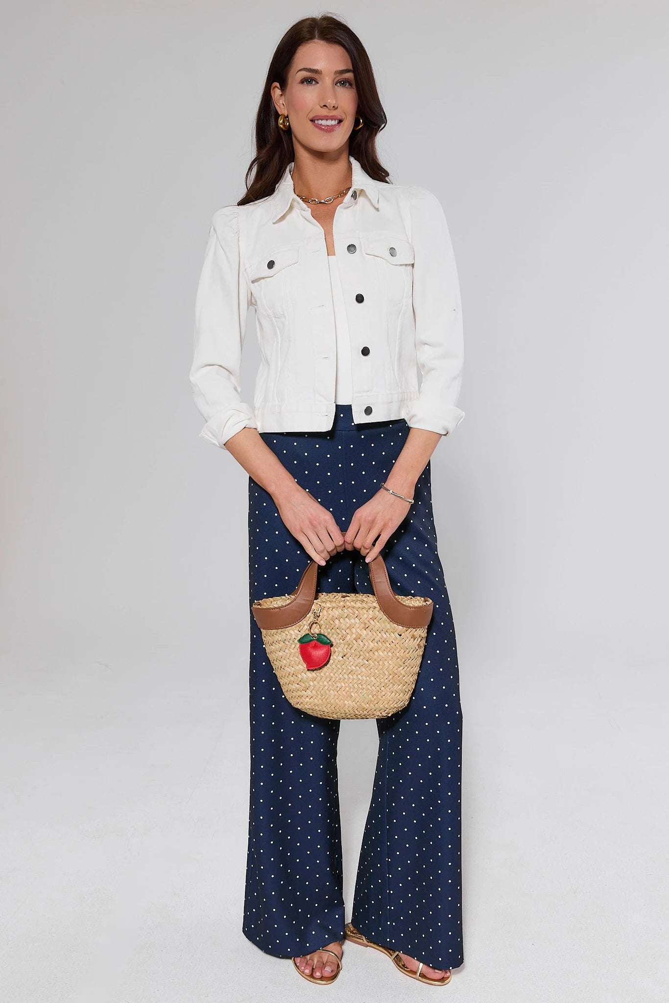 Woman holding a straw bag with an apple design, wearing a white jacket and navy polka dot pants on a plain background