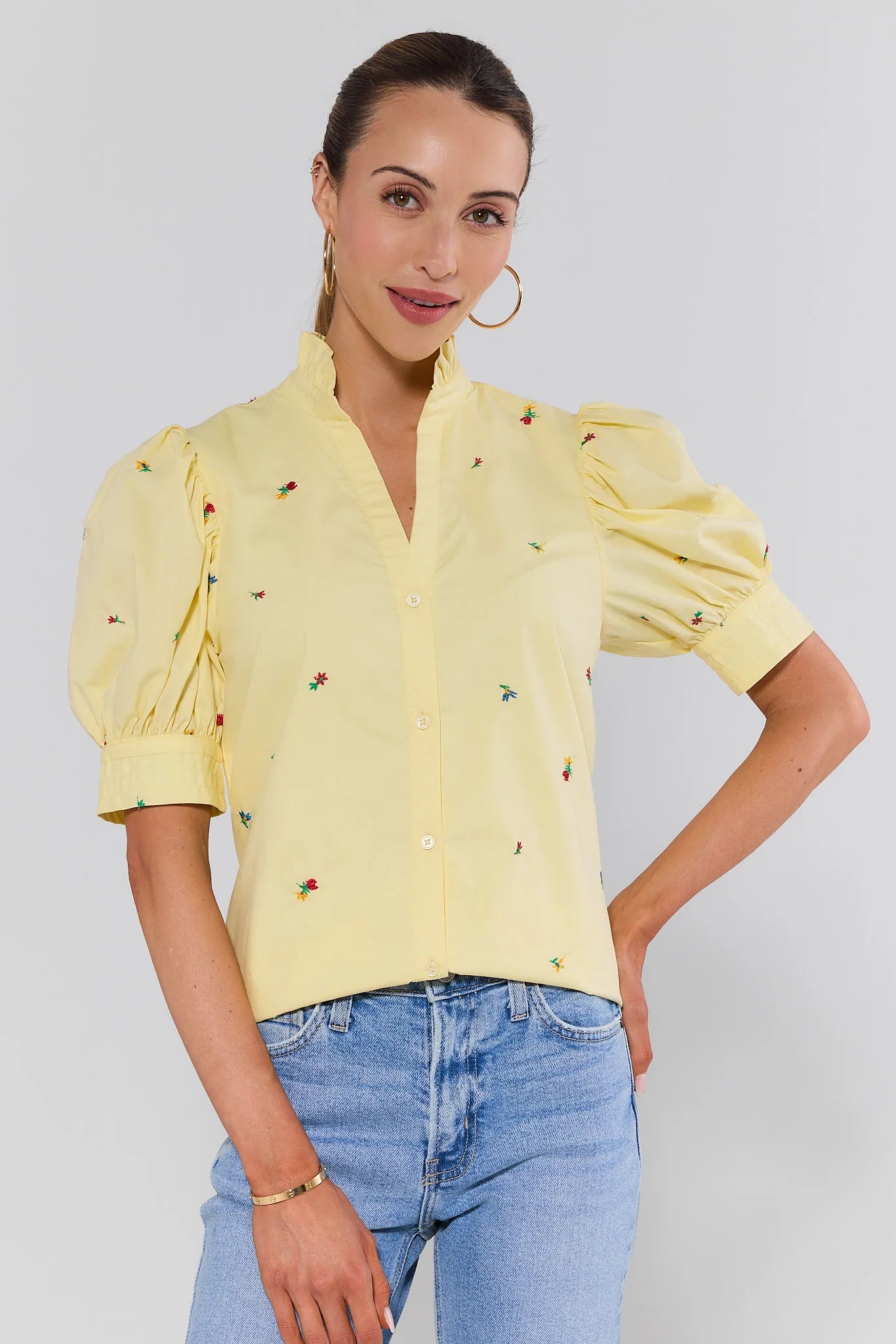 Woman wearing a yellow blouse with floral embroidery and blue jeans on a plain background