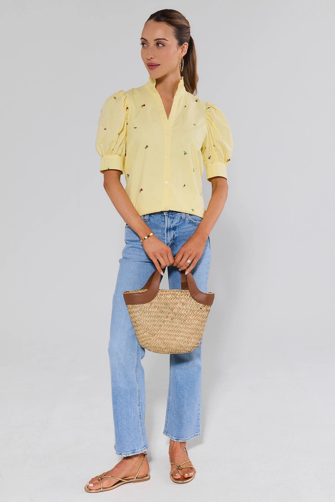 Woman wearing a yellow blouse and blue jeans, holding a straw bag on a plain background
