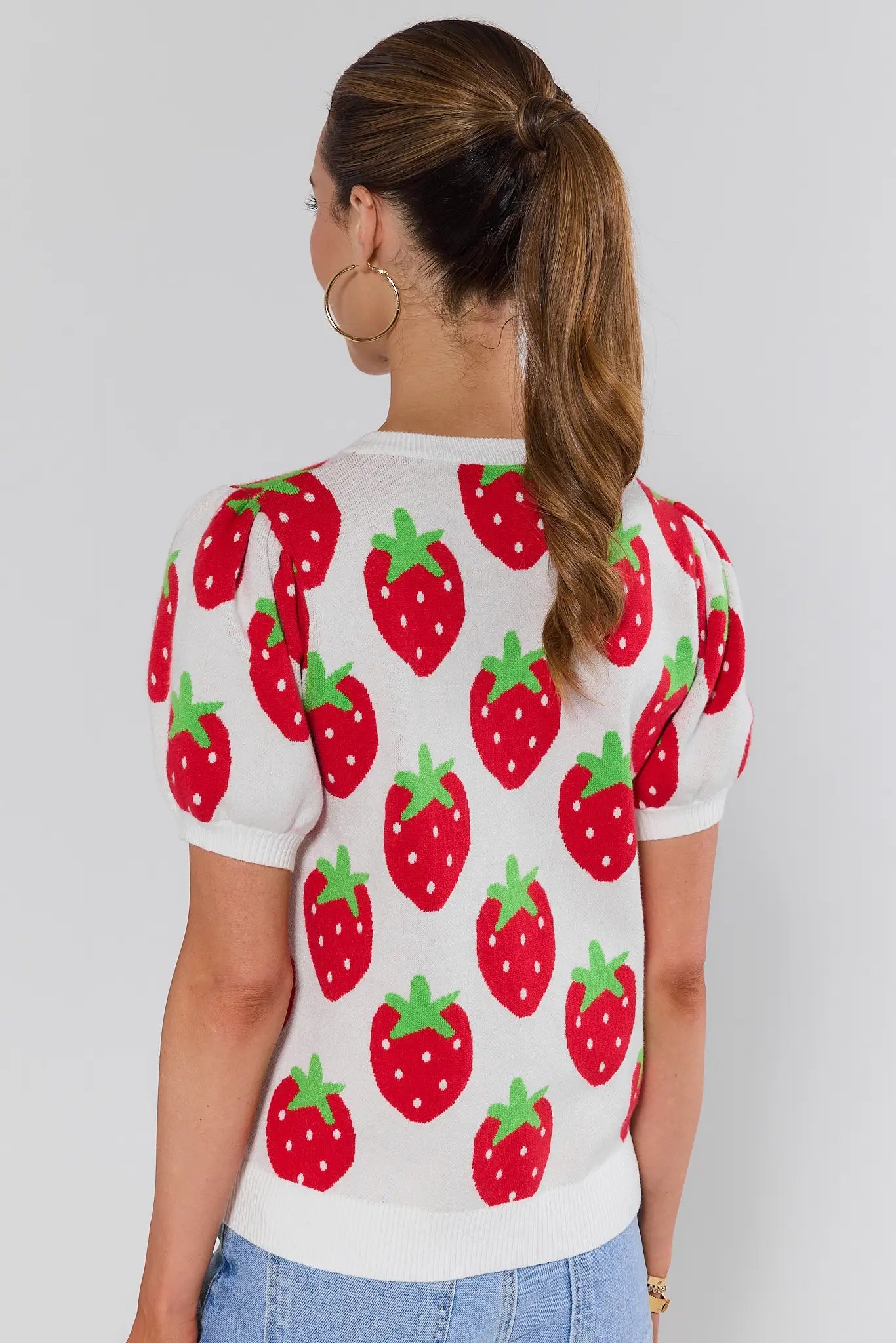 Person standing wearing a white sweater with red strawberry pattern on a plain background