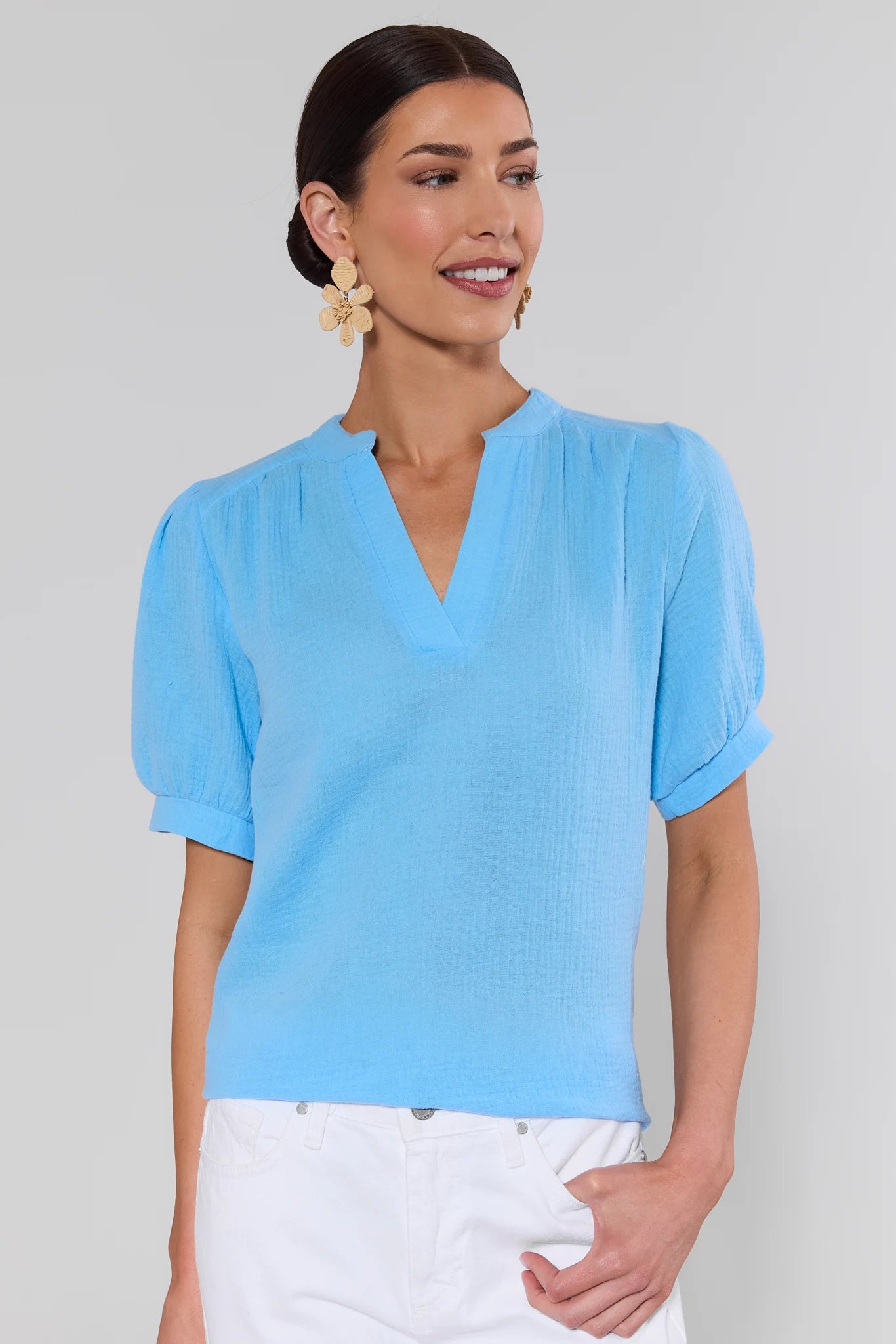 Woman wearing a light blue blouse with white pants and earrings against a plain background