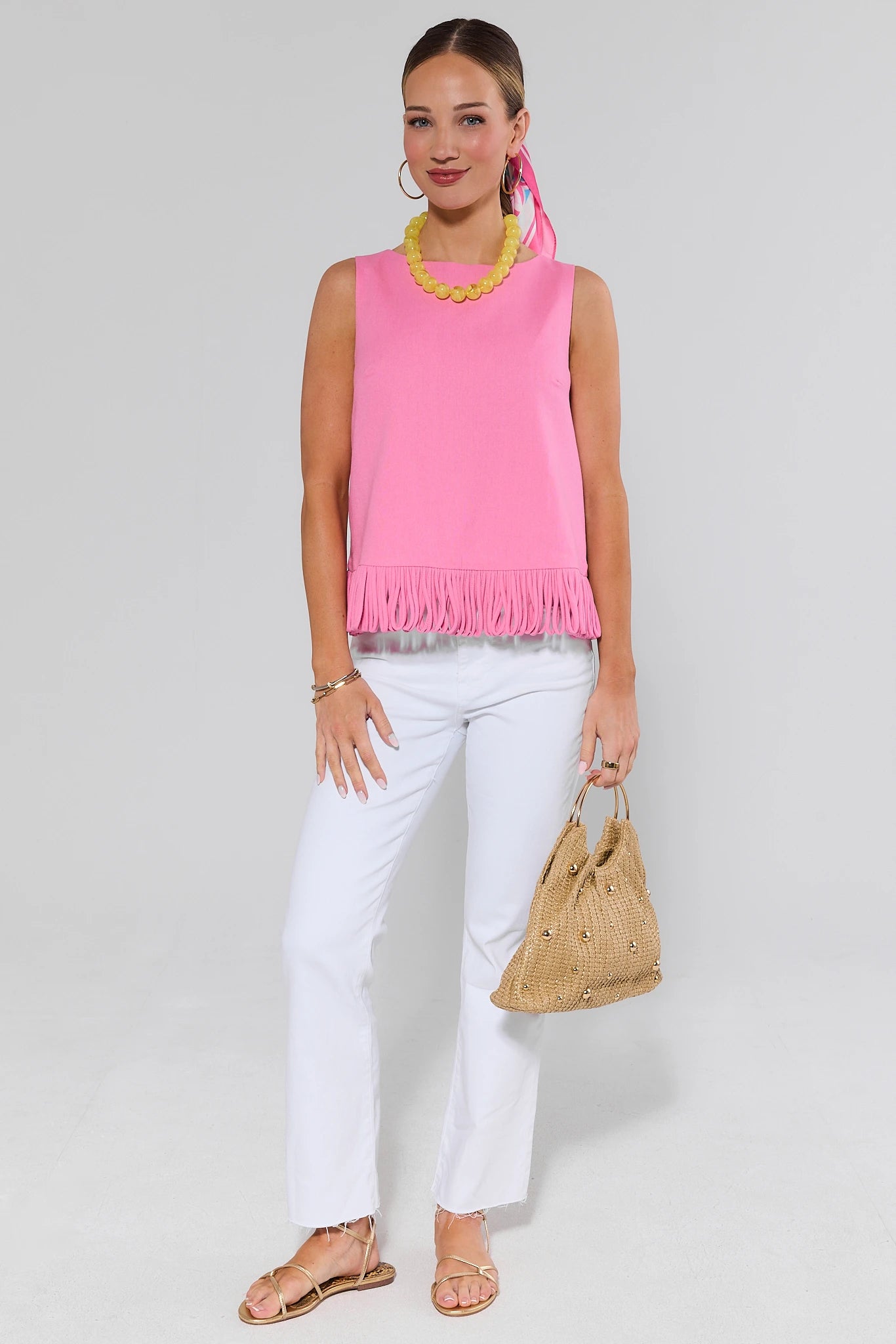 Woman wearing a pink sleeveless top and white pants, holding a straw bag on a plain background