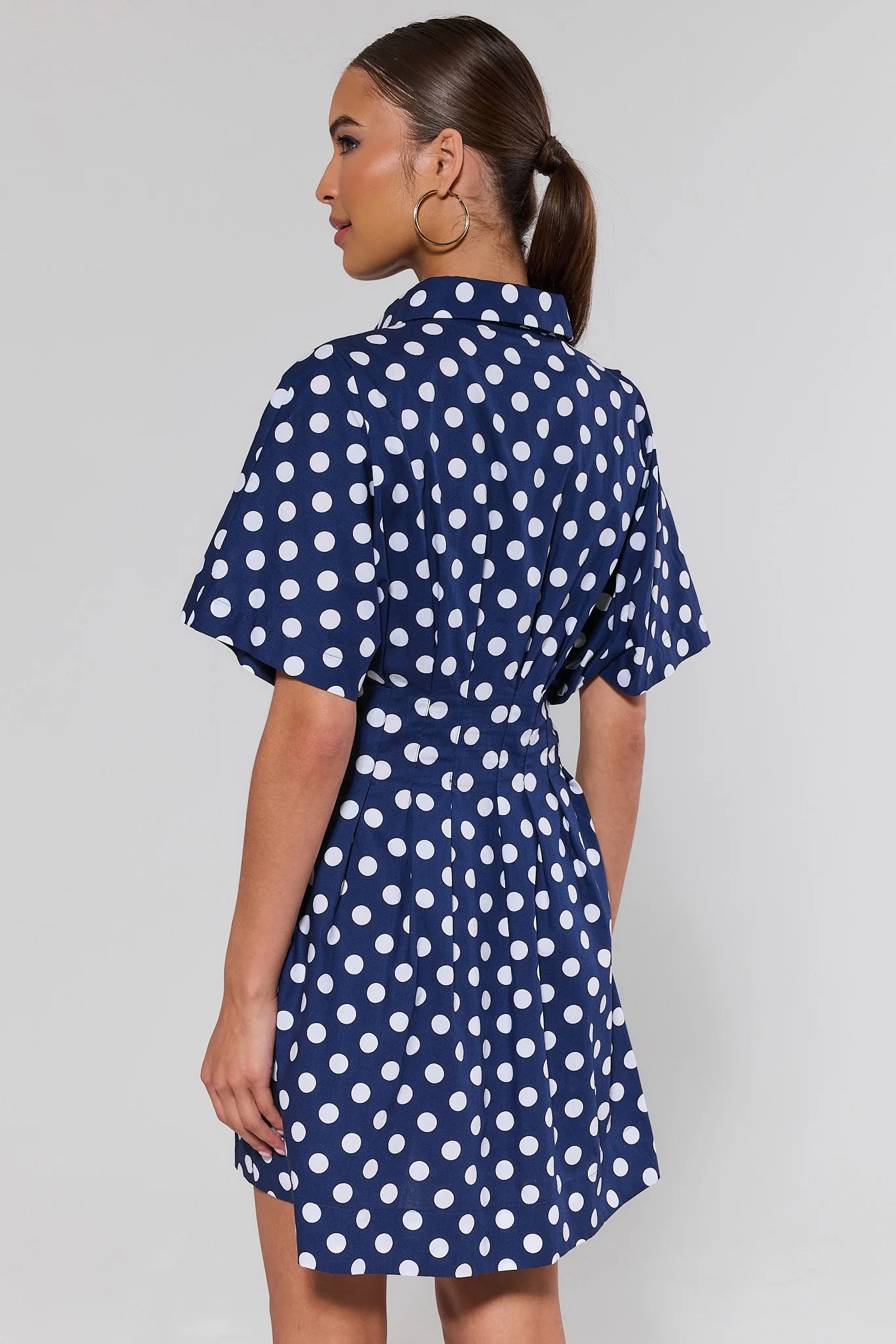 Woman standing wearing a navy blue dress with white polka dots on a plain background