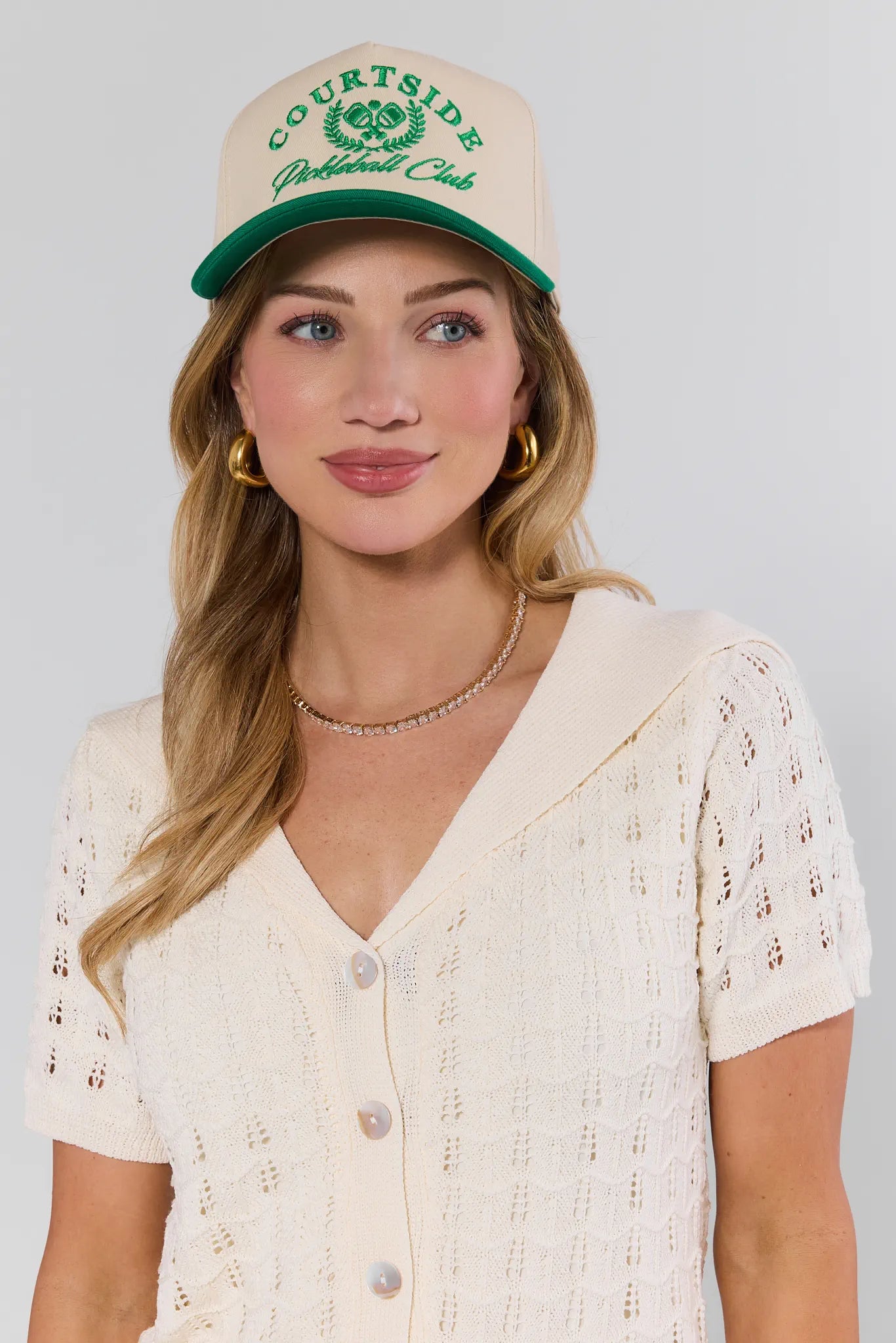 Woman wearing a beige cap with green text and a white lace top against a plain background