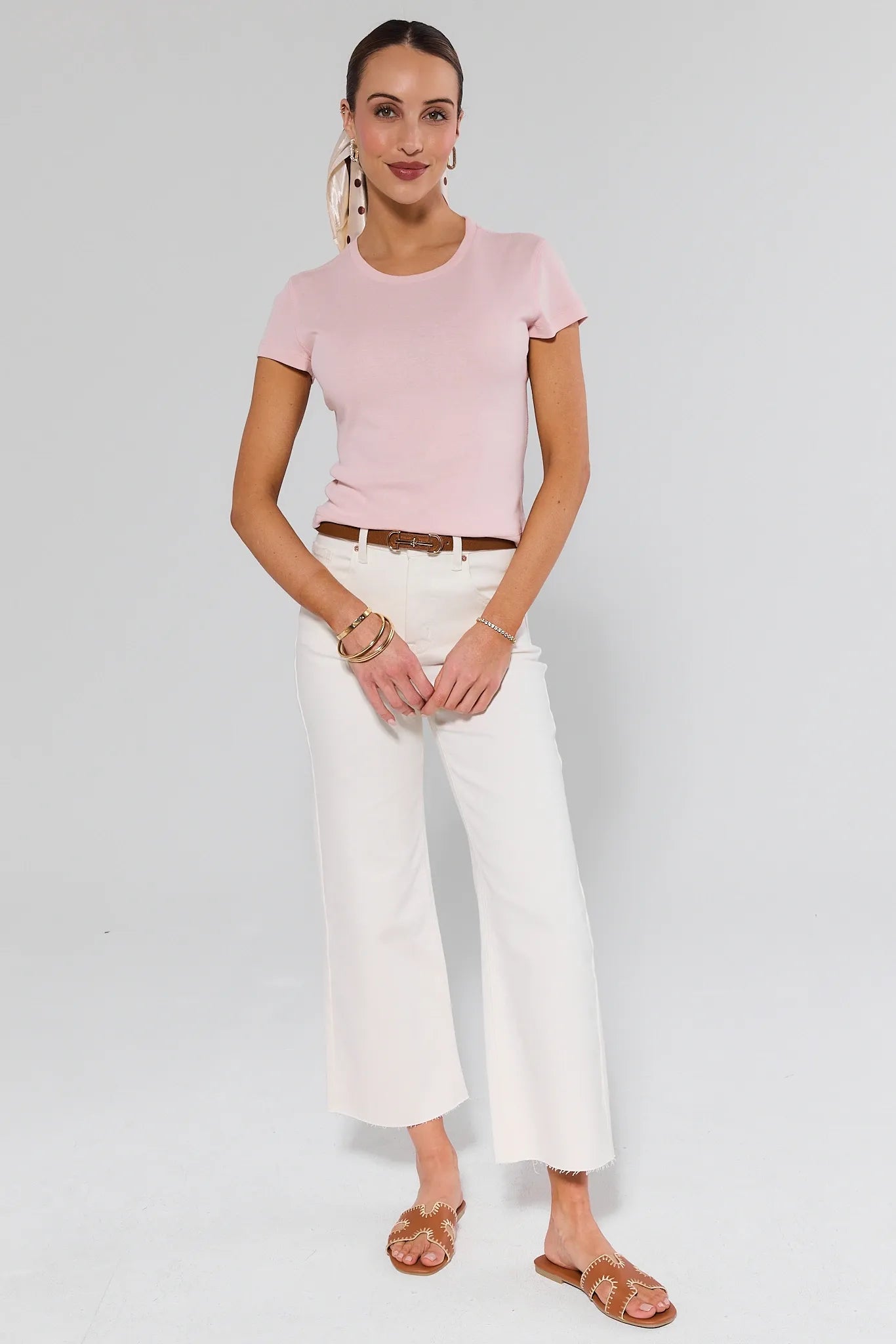 Woman wearing Caroline Ecru Straight Leg Jeans, styled with a pale-pink t-shirt, brown belt, tan slide sandals, and gold accessories, standing against a plain backdrop.