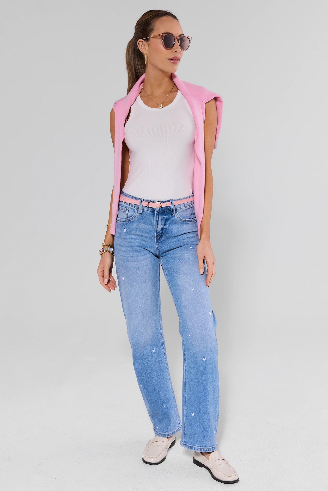 Woman wearing a pink sleeveless jacket, white top, blue jeans, and sunglasses on a light gray background