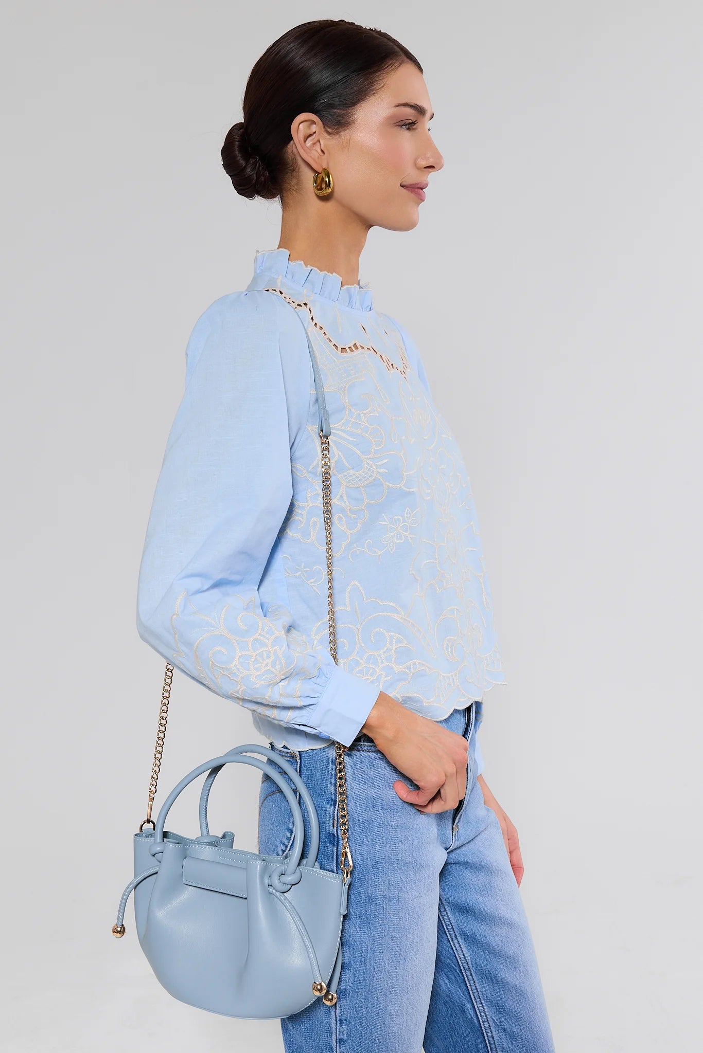 Woman wearing a light blue blouse with lace details and holding a matching handbag against a plain background