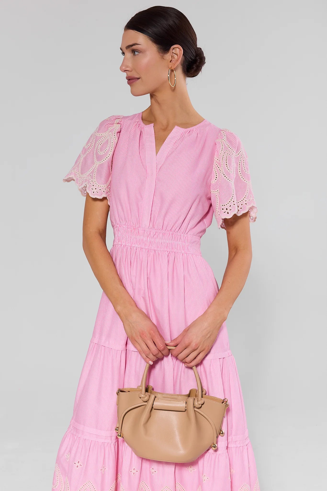 Woman wearing a pink dress with lace details holding a beige handbag against a light gray background
