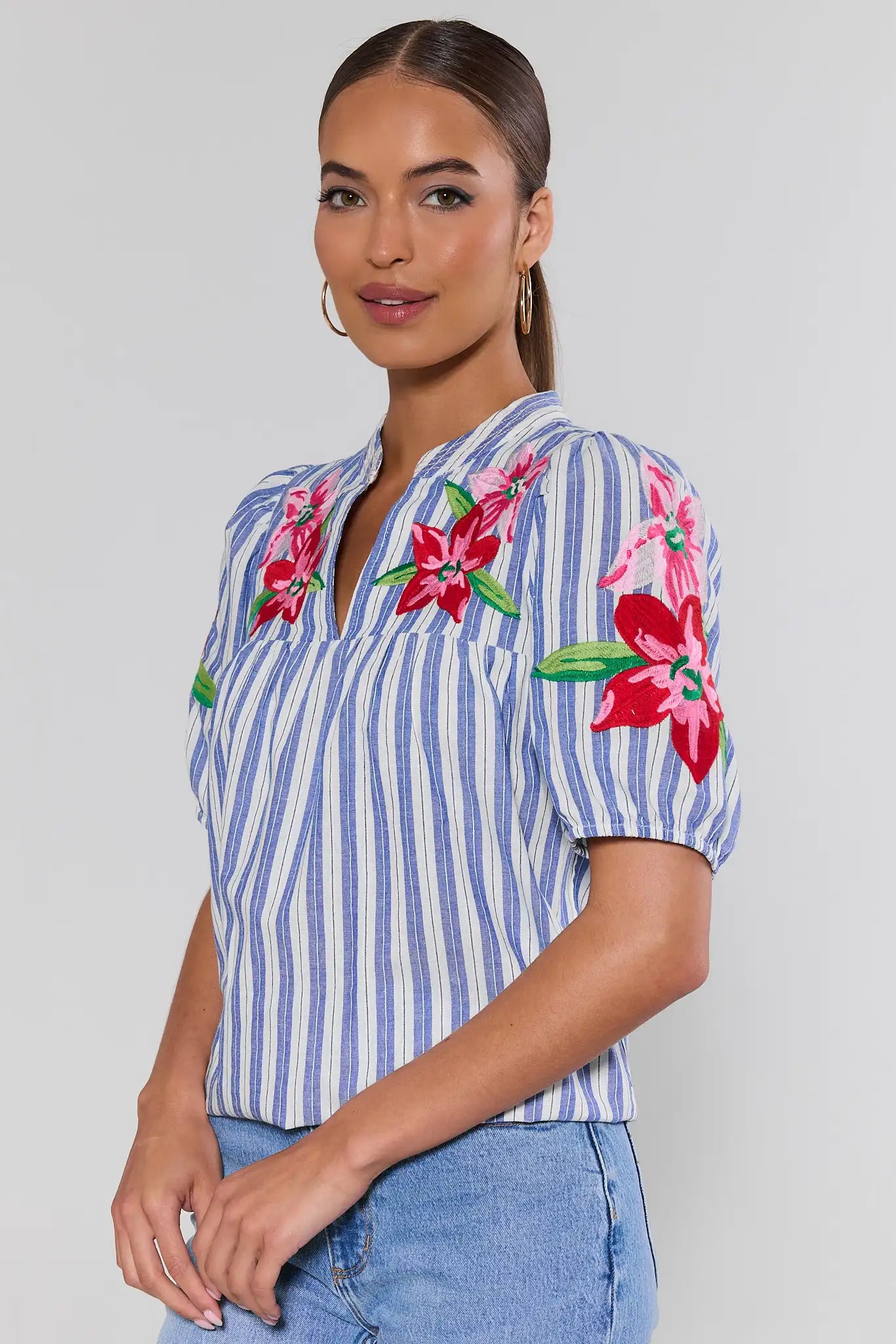Woman wearing a blue and white striped shirt with floral embroidery on a plain background side shot